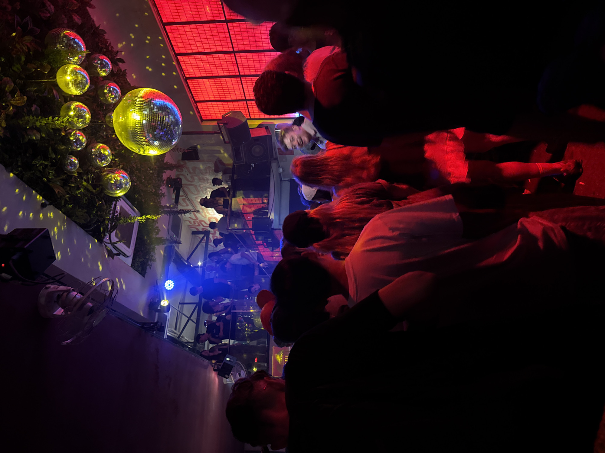 The image captures a lively scene in an unknown city. A group of people, mostly out of focus, are gathered in an outdoor venue illuminated with a mix of red, blue, and yellow lighting. Disco balls of various sizes hang amidst foliage on the left side of the frame, reflecting light in a scattered pattern. Overhead, a grid-like structure emits red light. A fan is mounted on the wall. The overall atmosphere suggests a party or event.