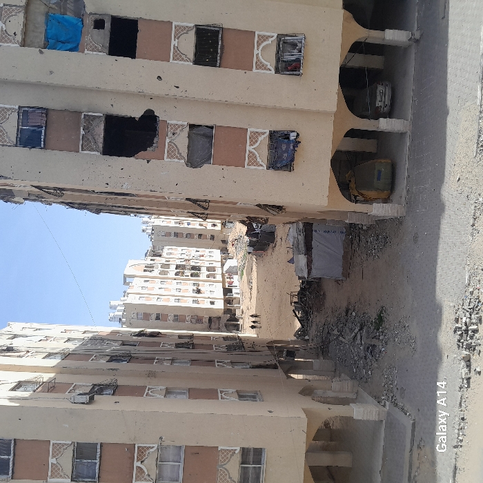War-torn apartment buildings flank a debris-strewn alleyway. Buildings have missing windows and visible damage. An excavator sits among the rubble. In the distance, other buildings line the horizon under a blue sky. The scene reflects devastation and destruction.