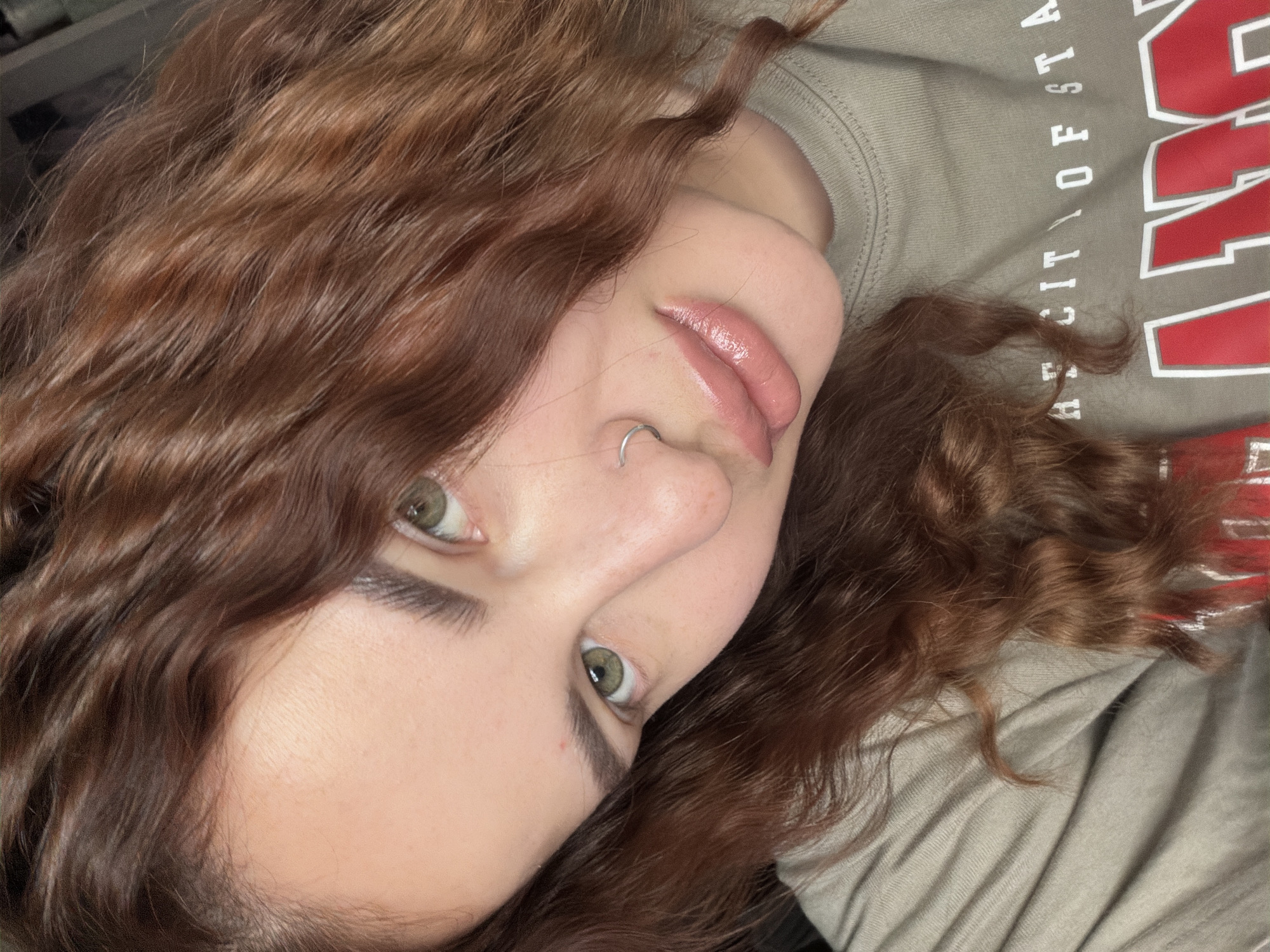 A close-up selfie of a young woman with wavy auburn hair. She has fair skin, green eyes, and a silver nose ring. Her lips are glossy. She is wearing a gray t-shirt with a red and white design that includes the words "CITY OF STA". The woman is the primary subject; there are no other people visible. The location is impossible to determine based on the image, as no landmarks or contextual clues are present, and the location in Novopokrovskaya, Russia, is not verifiable from the image itself.