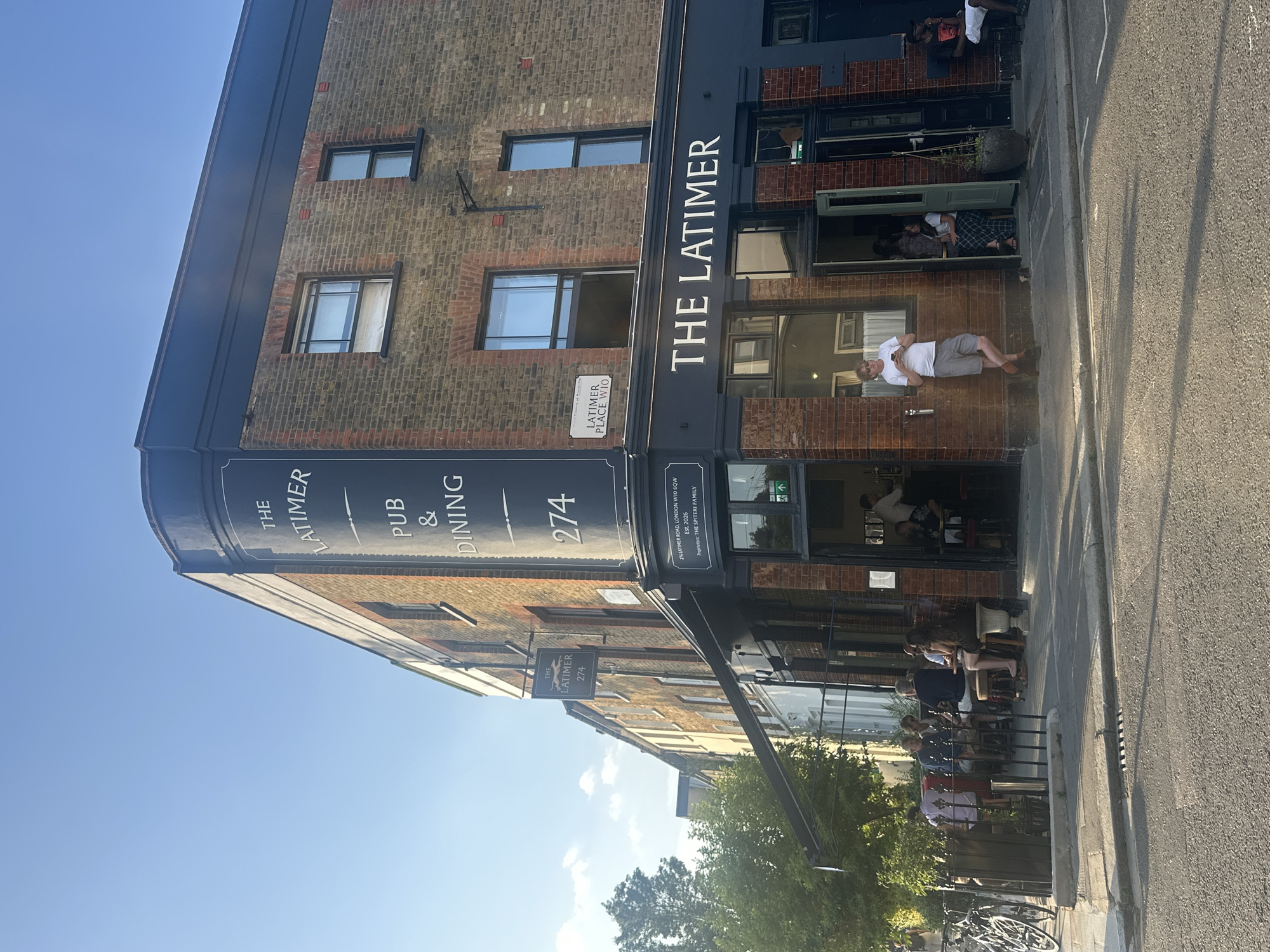 The image shows the exterior of The Latimer pub and dining establishment in London, United Kingdom. The building is a brick structure with a distinctive corner design featuring a prominent sign displaying "THE LATIMER PUB & DINING 274." A smaller sign indicates "LATIMER PLACE, W10" and "274 Latimer Road." People are seated outside at tables near the street, and others are standing or sitting inside the pub near the entrance. A bicycle rack with bicycles is visible near the sidewalk. The weather appears to be sunny.