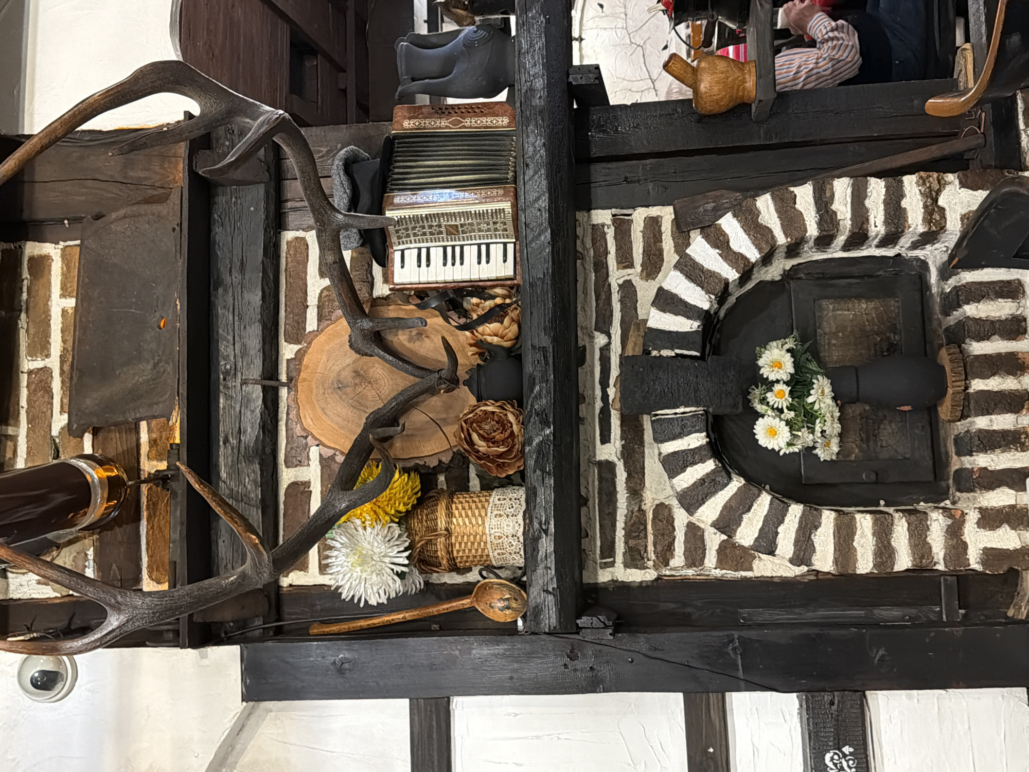 The image shows an indoor, overhead shot of a wall display. The wall is constructed of black and white half-timbered sections and brickwork. A pair of antlers is prominently displayed on the left side. Between the antlers and the right side of the frame, there is an accordion, a wooden log section, flowers, a basket with lace, and a wooden spoon. A person is visible in the background in the upper right corner of the frame. The right side of the frame contains a fireplace-like section with a black arched door, decorated with white daisies. The objects are arranged as decorations or art pieces. There is a security camera mounted below the antler display. Based on search results, the scene is located in Varna, Bulgaria.