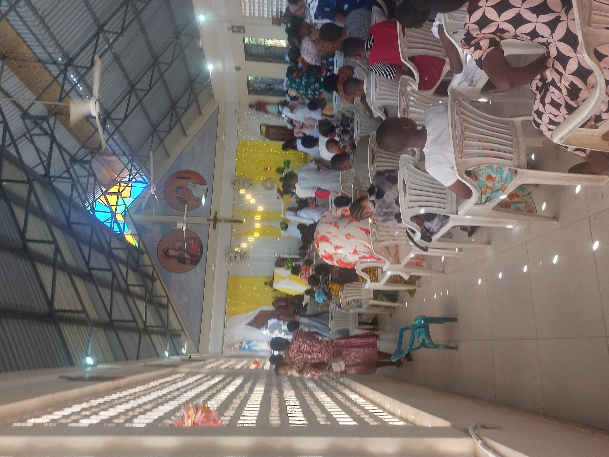 The image shows an indoor scene in what appears to be a church. People are seated on white plastic chairs, facing forward. At the front, there is a decorated altar area with yellow and white fabric. Wall decorations include circular portraits and a cross. A stained glass window is visible. Ceiling fans are present. The building has a metal roof structure. Considering the presence of people and the structure, the location is likely a church setting in Dar es Salaam, Tanzania.