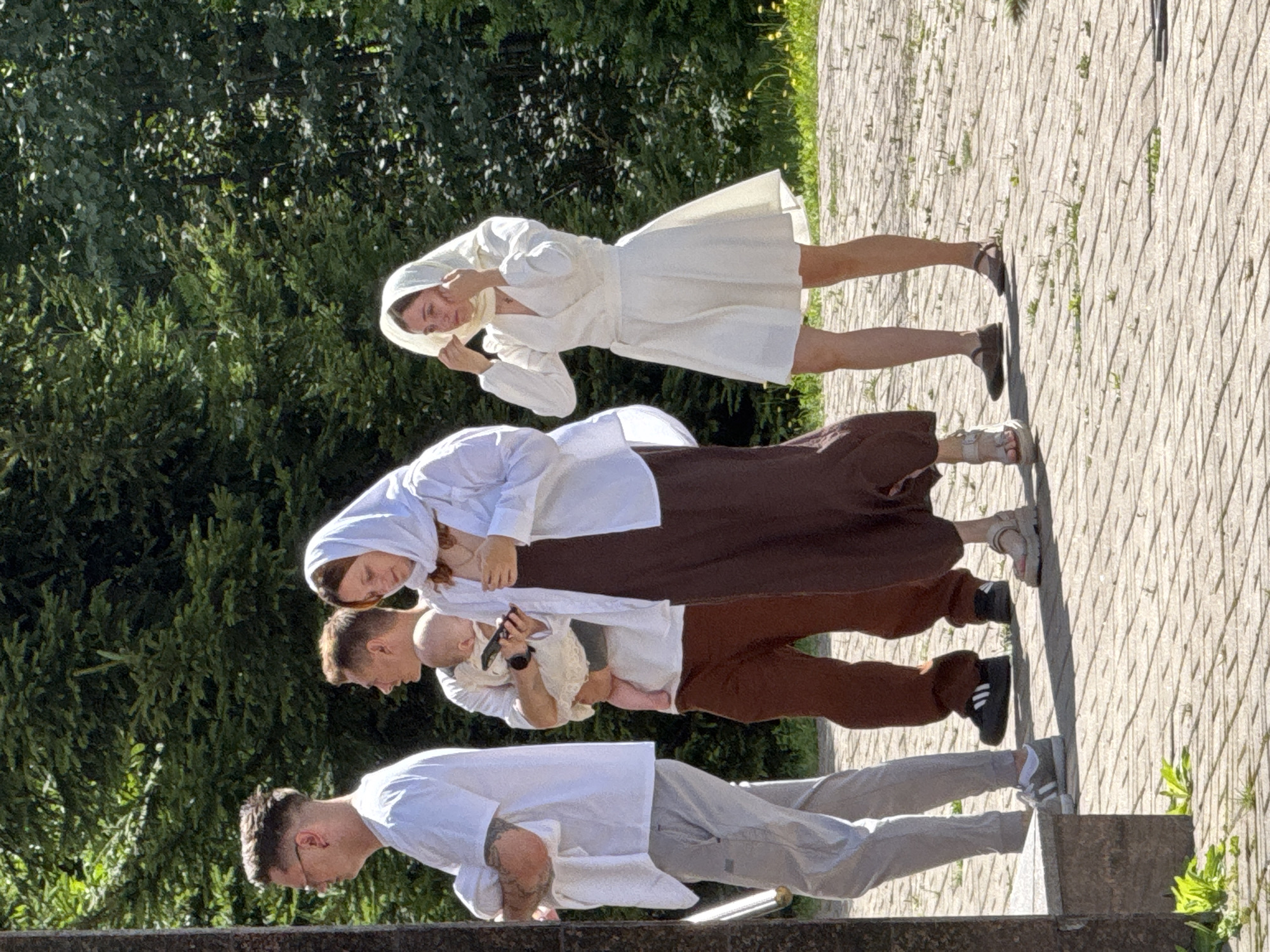 The image shows four people walking on a paved pathway lined with trees. A man on the left, wearing glasses and a white t-shirt, holds a baby. Next to him, a man in a white t-shirt carries a baby. A woman in a brown dress and white headscarf walks behind them, holding a mobile phone. On the right, a woman in a white dress and headscarf adjusts her headscarf. The background consists of dense green trees. The pathway appears to be made of light-colored brick or stone. The lighting suggests it is daytime, with sunlight casting shadows. Based on the architectural style of the pathway and the presence of lush greenery, this scene could be located in a park or public garden within Saint Petersburg, Russia, possibly near a church or other religious institution given the attire of some individuals.