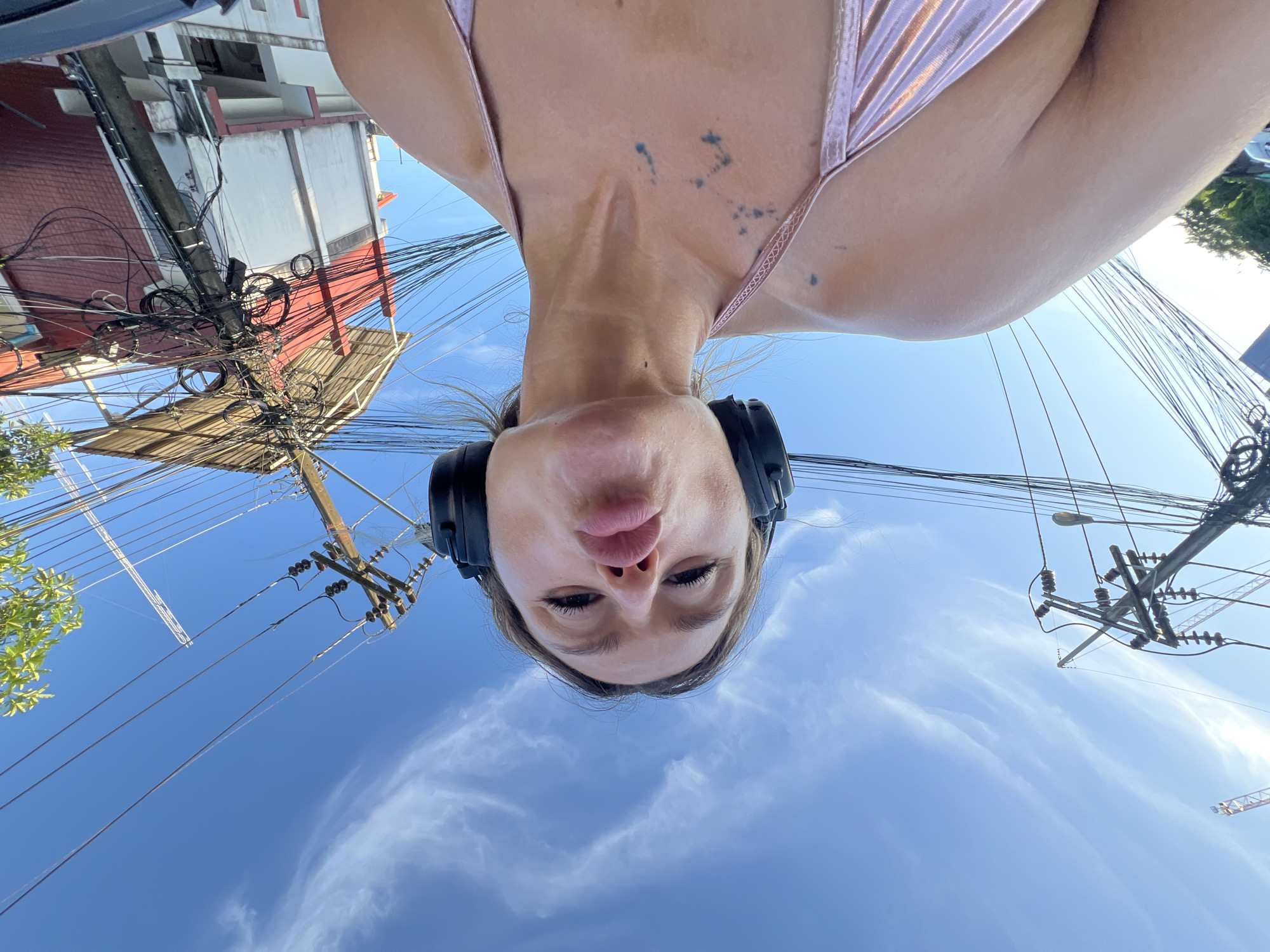 A low-angle, wide-angle selfie shows a woman wearing black headphones and a metallic pink bikini top, puckering her lips at the camera. She has a small, abstract tattoo on her chest. The background is a bright blue sky with wispy white clouds. Overhead, a dense network of power lines and utility poles are visible, with one pole prominently displaying a tangled mass of wires and cables. A red and white building with a covered balcony is partially visible behind the utility pole, suggesting an urban or semi-urban setting. The scene appears to be outdoors, possibly on a street in Choeng Thale, Thailand, given the characteristic dense electrical wiring commonly found in Southeast Asia.
