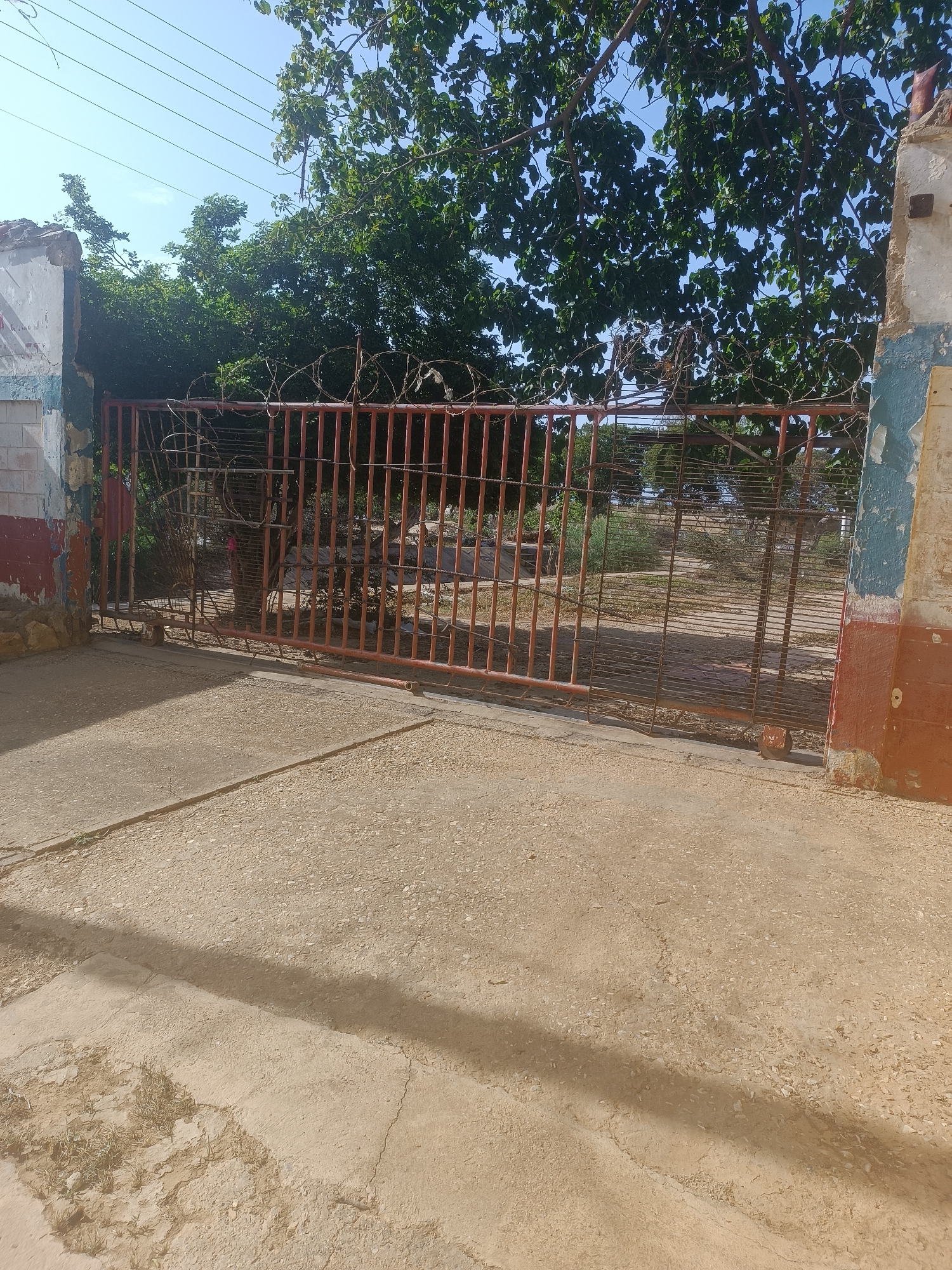 A ground-level, medium shot shows a fenced entrance on a sunny day. The fence is a rusty red metal gate with vertical bars, topped with barbed wire. The gate is partially open, allowing a view to a grassy area beyond. The entrance is flanked by two decaying concrete structures, one on each side. The structures are partially painted with red, white, and blue sections. The ground is a cracked, light-brown concrete surface. Green trees are visible above and behind the fence. The location appears derelict.