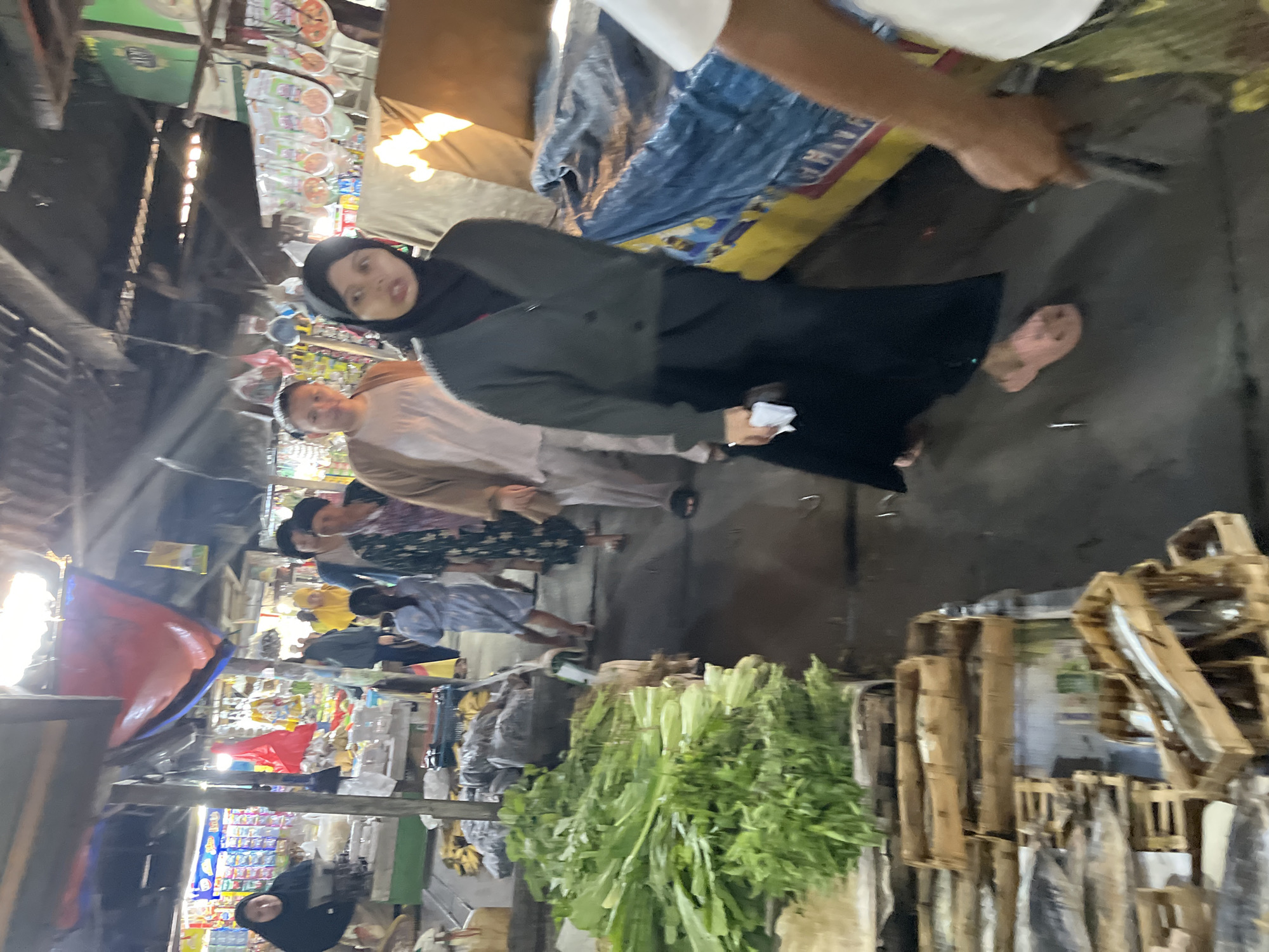 The image depicts a bustling outdoor market scene in Larangan, Indonesia. A woman wearing a black hijab and a dark green cardigan walks on the right side of the frame, holding a white cloth in her left hand. She is wearing pink sandals. To her left, a person in a light brown cardigan and white top is visible, partially obscured. Further left, a woman in a patterned green and white dress walks away from the camera, and another woman in a beige outfit is behind her. In the foreground, to the left, a large pile of fresh green vegetables is displayed on a wooden surface. Behind the vegetables, wooden crates containing what appear to be fish are arranged. The market environment features a covered walkway with a corrugated metal roof, strung with various items. The ground is wet, suggesting it might have rained or been recently cleaned. The overall scene conveys the lively atmosphere of a traditional Indonesian market.