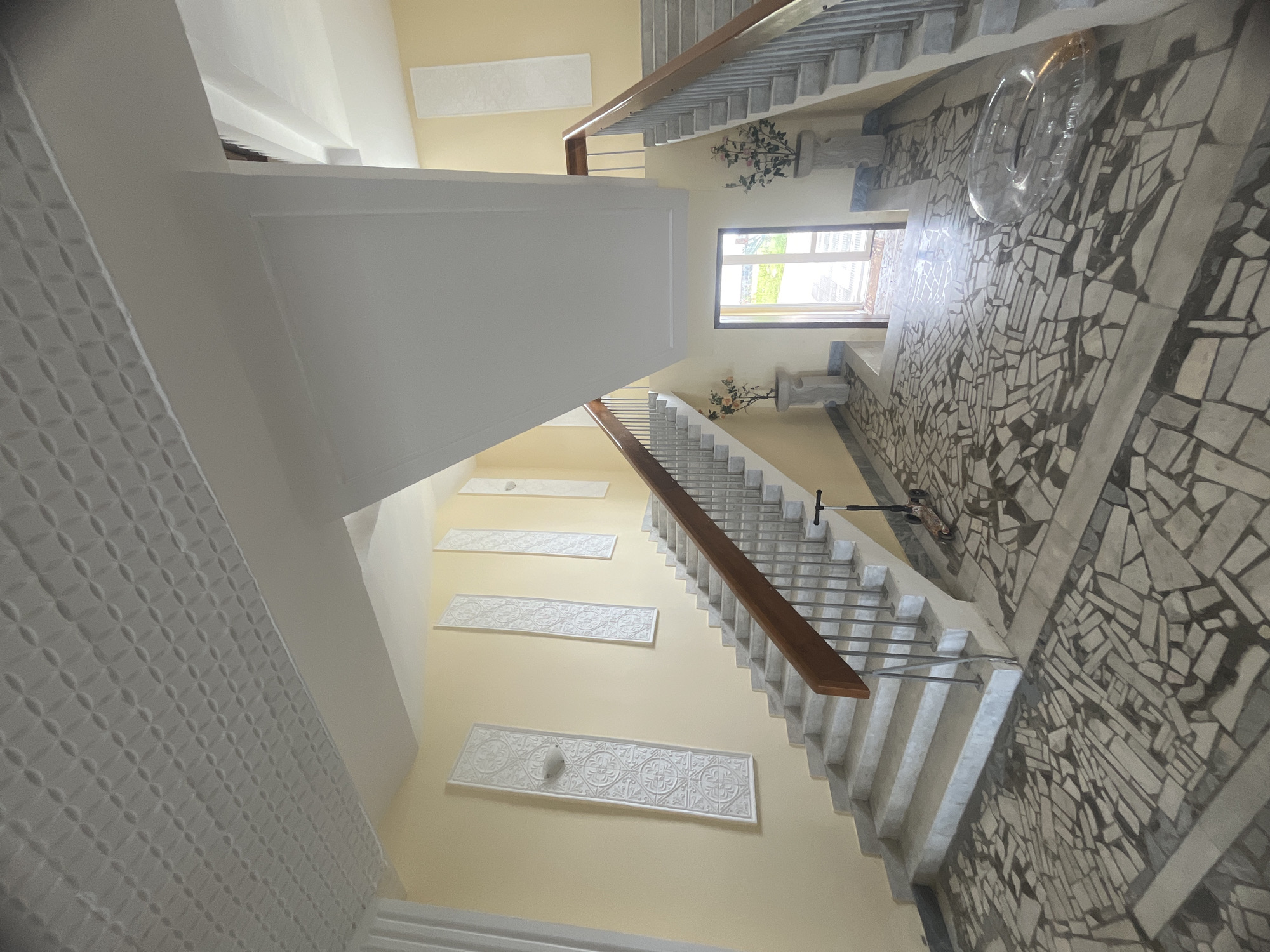 This is a wide-angle photograph of an interior staircase and landing area within a building. The walls are painted a pale yellow, and the ceiling features a textured, patterned design. Ornate, rectangular white panels are mounted vertically on the walls. The staircase itself has marble steps and a wooden handrail supported by a metal railing. The flooring is a mosaic of irregular, light-colored tiles.
To the left, a scooter is parked on the floor near the bottom of the staircase. On the landing in the center of the image, there is a doorway leading outside, with light filtering through. Two decorative planters with artificial flowers are positioned on either side of the doorway. In the foreground on the right, a transparent inflatable ring lies on the mosaic floor.
The scene suggests a residential or public building's interior, possibly an apartment building or a hotel, given the architectural style and the presence of decorative elements. The lighting appears to be natural, coming from the exterior doorway and an overhead source.
The image is undated and unsigned, but the style of the architecture and decorative elements could suggest it was taken in the mid to late 20th century.
While I can describe the visual elements of the image, I cannot determine the specific location within the city of Sokhumi, Abkhazia, based solely on the provided visual information.