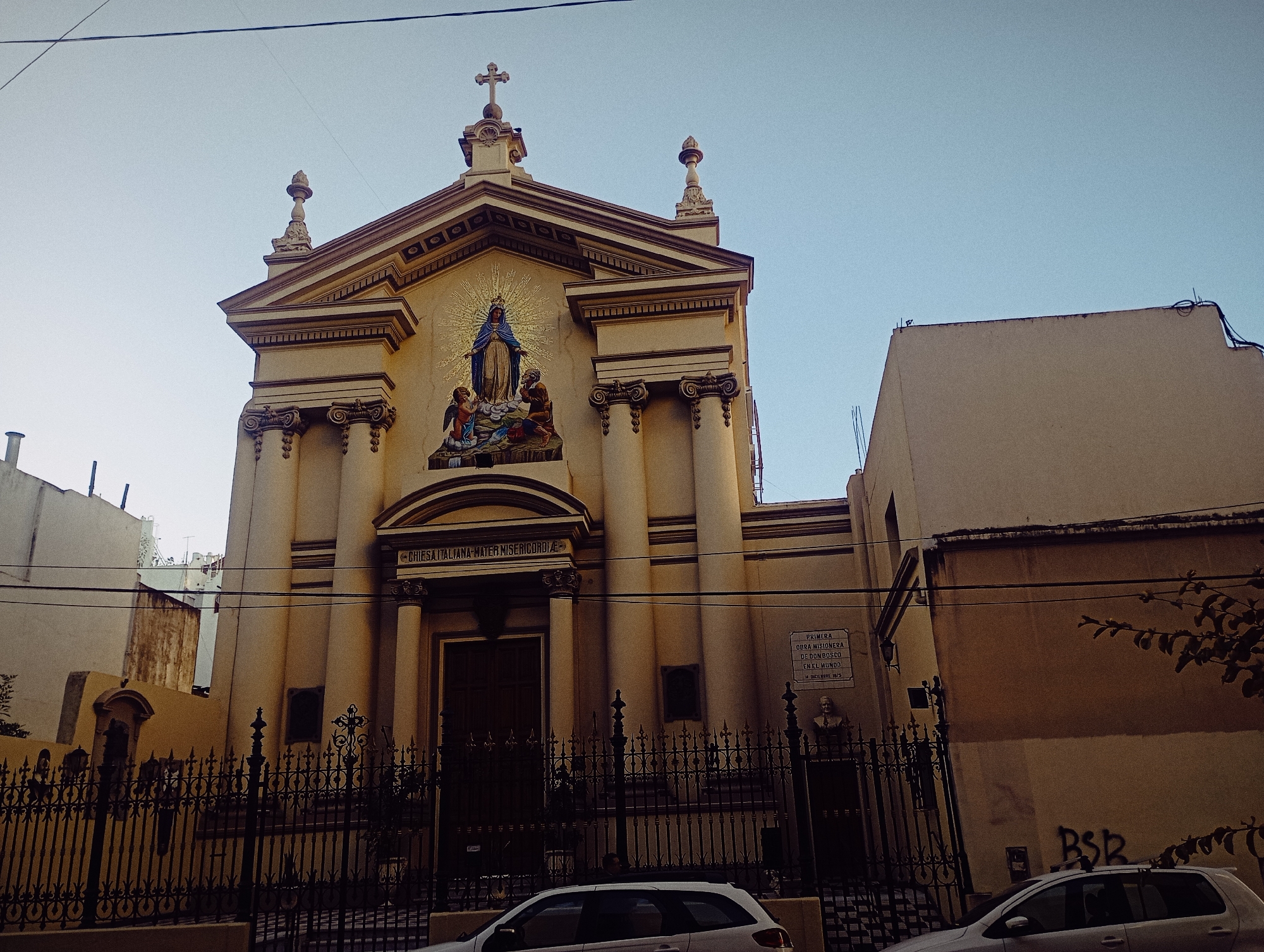 The image presents a low-angle view of the Chiesa Italiana Mater Misericordiae (Italian Church of Mother of Mercy), located in Buenos Aires, Argentina. The church facade is a light yellow color, with a classical architectural style featuring columns, pilasters, and a pediment. Above the entrance, there is a mosaic depicting the Virgin Mary with figures kneeling before her. A wrought iron fence encloses the church grounds. A plaque on the side of the church reads "PRIMERA OBRA MISIONERA / DE DON BOSCO / EN EL MUNDO / 14 DICIEMBRE 1875," indicating the church's historical significance as Don Bosco's first missionary work in the world. A bust is situated next to the plaque. The sky is light blue and clear. Two white cars are parked along the street in front of the church, and adjacent buildings are visible on either side. There is graffiti on the side of one of the buildings. Overhead power lines are visible.