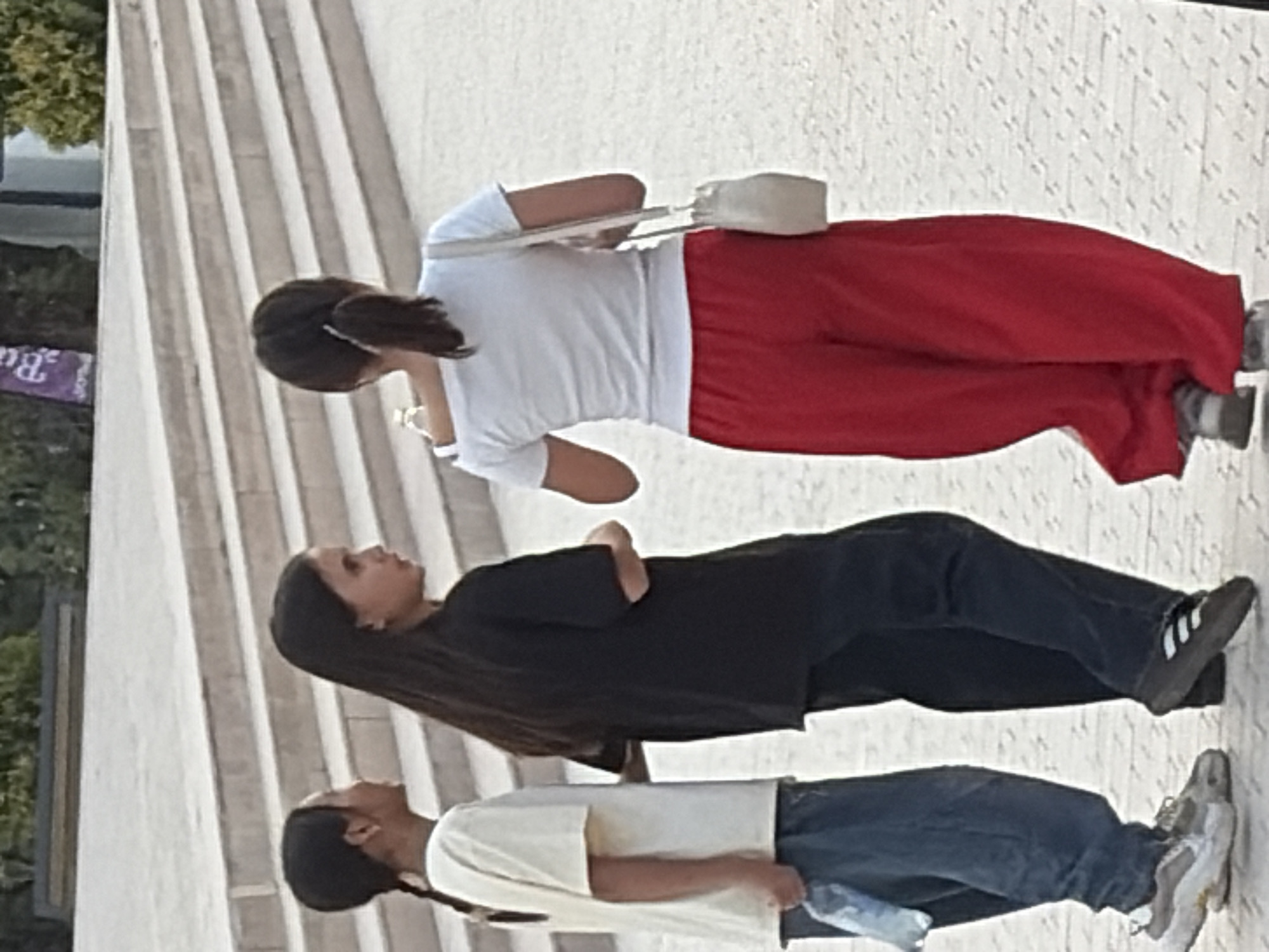 Three young females are walking away from the camera on a paved surface. The female on the left wears a light-colored t-shirt and blue pants, holding a plastic bottle in her left hand. The middle female is dressed in a black t-shirt and black pants. The female on the right wears a white off-the-shoulder t-shirt and red pants, carrying a light-colored shoulder bag. Behind them are concrete stairs and some greenery. The background shows a purple banner with white lettering.