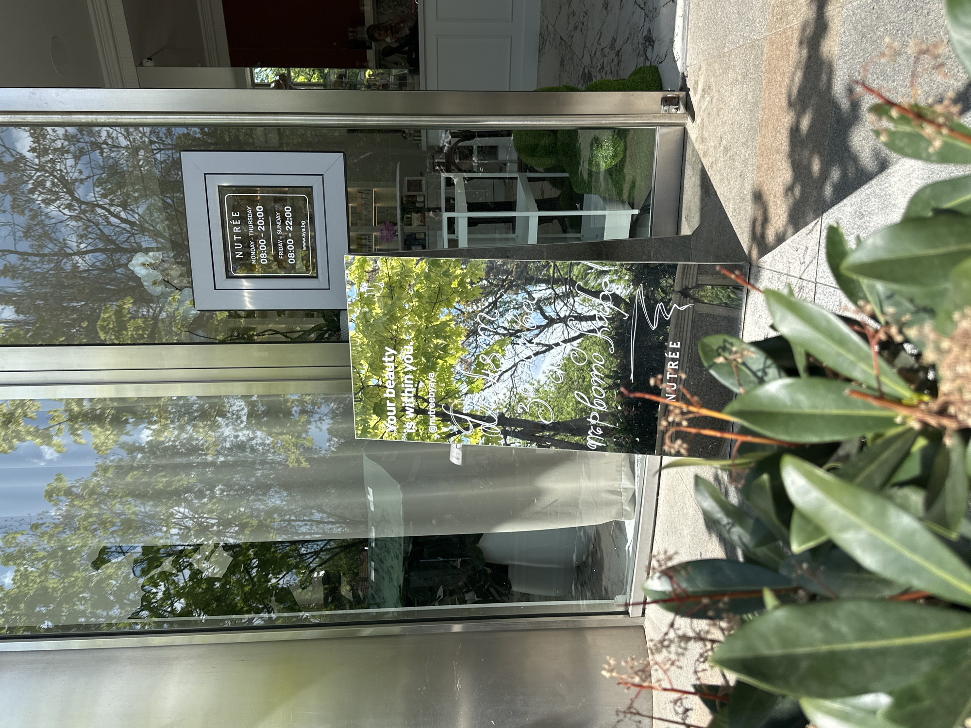 A storefront of "NUTRÉE" in Sofia, Bulgaria is presented in a daytime exterior shot. A sign affixed to the glass indicates operating hours: Monday to Thursday, 08:00 to 20:00, and Friday to Sunday, 08:00 to 22:00, with the website "www.aya.bg" listed. A promotional sign is also visible, stating, "Your beauty is within you," with the Instagram handle "@nutreebyaya" displayed. The storefront's interior contains a visible white display shelf with cosmetic products on display. The entrance is flanked by greenery. The outside sidewalk is tiled, and the scene is cast in bright sunlight.