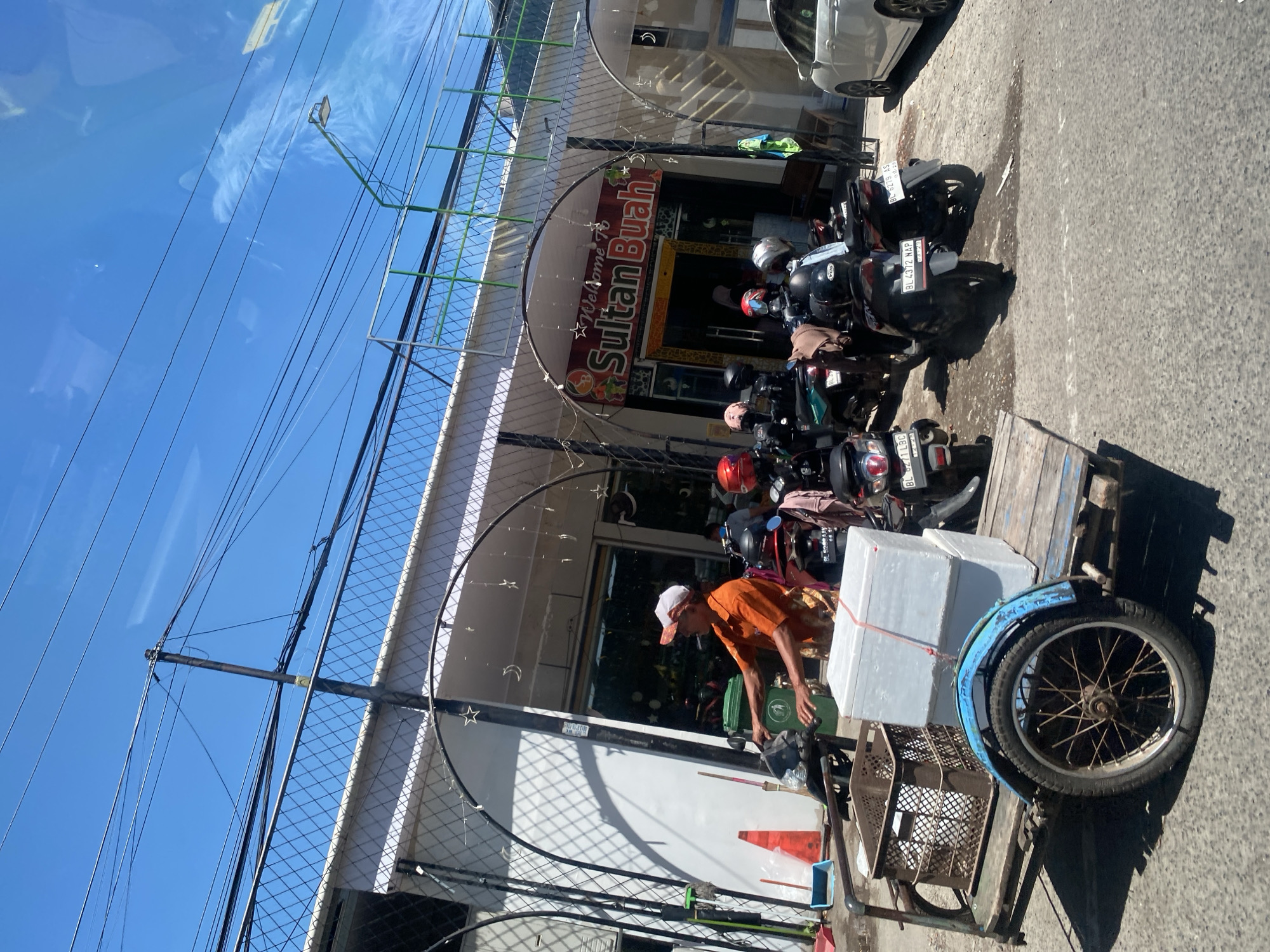 A man in an orange shirt and a white baseball cap is standing next to a three-wheeled trailer. The trailer is carrying a brown plastic basket filled with items and two large white boxes. The man appears to be operating a motorbike attached to the trailer. In the background, several motorbikes are parked in front of a building with a sign that reads "Welcome to Sultan Buah." The building has arched metal decorations and is decorated with string lights in the shapes of stars and moons. A white car is parked to the right of the building, and the sky is clear blue. The scene appears to be a street in Banda Aceh, Indonesia, on a sunny day.