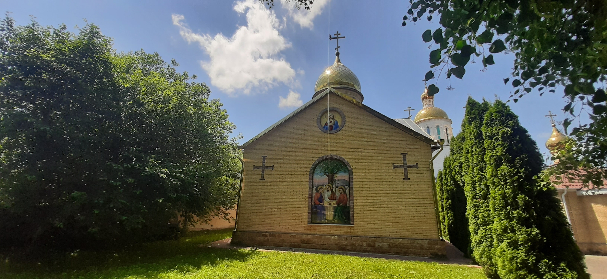 The image shows a yellow brick church with a golden dome against a bright blue sky with scattered white clouds. The church has a fresco above an arched window on its facade. The window itself also features a fresco, depicting three figures and a tree. Two crosses are visible on the exterior walls of the church. The building is surrounded by lush green trees and a well-maintained lawn. In the background, other golden-domed structures and evergreen trees can be seen. The scene suggests a serene, daytime setting in a religious complex.