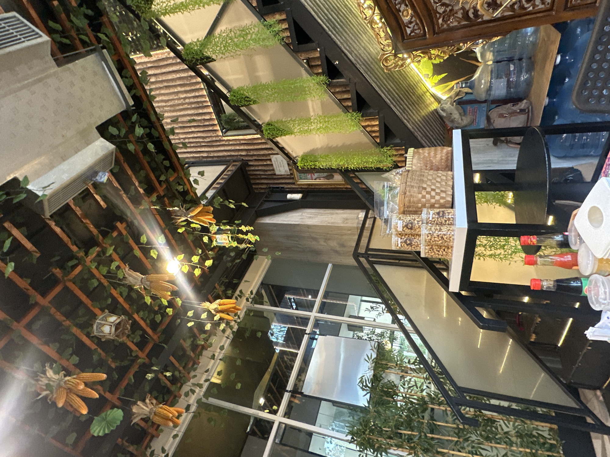 The image depicts an indoor space in Central Jakarta, Indonesia. The architectural style is modern with an emphasis on natural light and greenery. There are large windows, and decorations include plants, artificial corn, and other foliage. The layout suggests a commercial establishment, possibly a restaurant or cafe. Furniture is visible, including a table and chairs. Shelves are stocked with goods, and condiments are placed on a table. The overall atmosphere is a blend of contemporary design and natural elements.