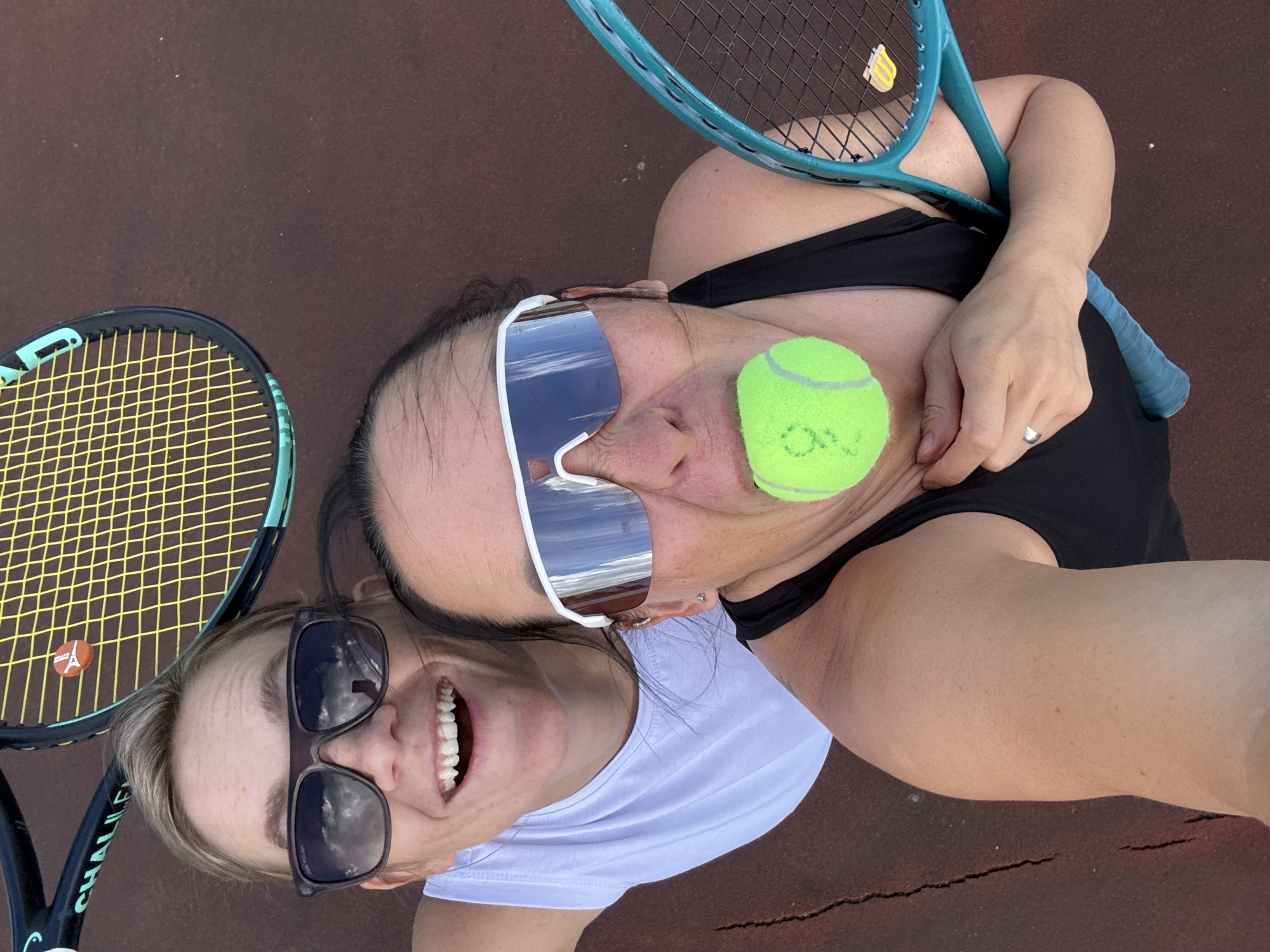 Two women are on a tennis court, one in the foreground and one behind her. The woman in the foreground is wearing a black tank top and large white wrap-around sunglasses, with a tennis ball in her mouth. The woman behind her is wearing a white tank top and dark sunglasses. Two tennis rackets are visible, one with a yellow and black strung pattern and a small orange logo, and another with a teal frame and strings. The court surface is a reddish-brown color. The image appears to be a selfie taken from a high angle. The location of Chaikovsky, Russia, is not discernible from the image.