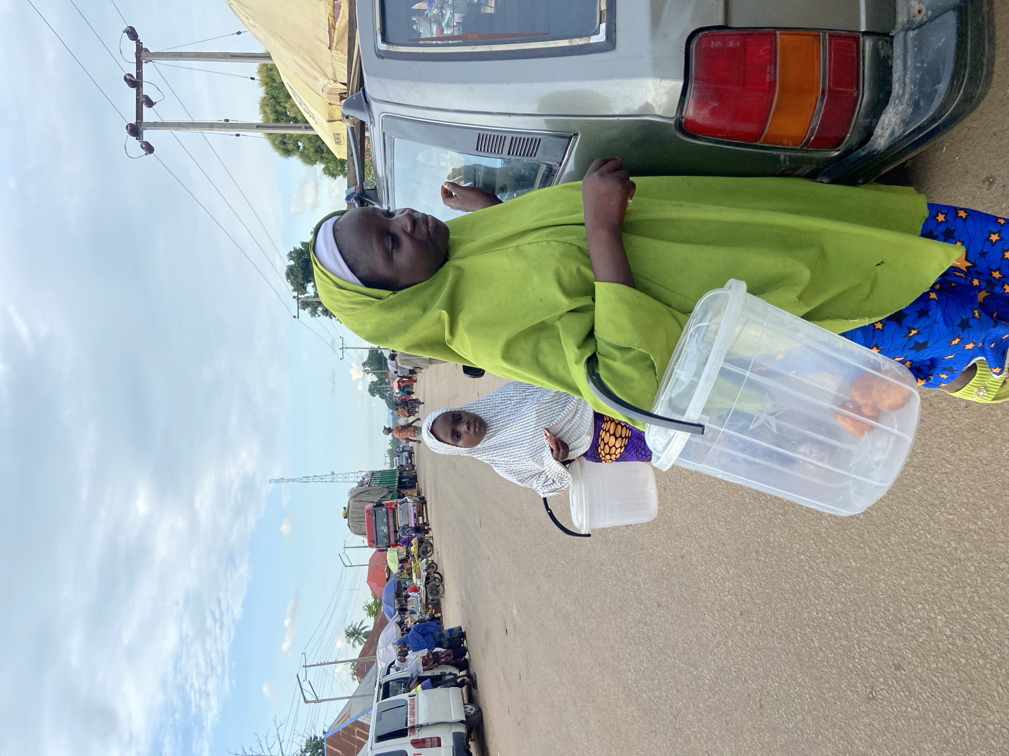 This image captures a daytime street scene in a busy commercial area of Mararaba, Nigeria, likely a market.
In the immediate foreground on the right, a woman with dark skin stands beside the rear passenger door of a dark-colored SUV or similar vehicle. She wears a lime green flowing garment over blue trousers adorned with yellow star patterns, and a white inner head covering beneath a matching lime green hijab. Her left hand holds a transparent plastic bucket with a black handle, containing several orange-colored items. Her right hand is positioned near the vehicle's window. Directly behind her, slightly to the left in the midground, stands a younger girl with dark skin, wearing a white patterned hijab, a white top, and a purple skirt with orange circular motifs. She holds a smaller transparent plastic bucket with a black handle, containing a light-colored, possibly rectangular, object. Both individuals face slightly towards the viewer, though the woman's gaze is directed downwards and to her right, while the girl looks more directly at the camera.
The road surface is paved with visible aggregate. Along the left side of the road in the midground, a white van marked "TAOS INT CORPORATION" is parked, with another white van partially visible behind it. Several individuals, primarily adults, are gathered around these vehicles and near market stalls with various colored umbrellas (red, blue) and beige canvas coverings. Fruit stands and other vendor carts are present. Further down the road on the right, a large red and green truck, heavily laden with goods covered by a tarpaulin, is visible, alongside other vehicles and more people. In the background, low-rise buildings with corrugated metal roofs are discernible on the left. Multiple wooden utility poles with overhead power lines span the scene, and a metal lattice telecommunication tower rises in the far background on the right. Sparse trees and foliage dot the horizon. The sky is partially cloudy with varying shades of blue and white.