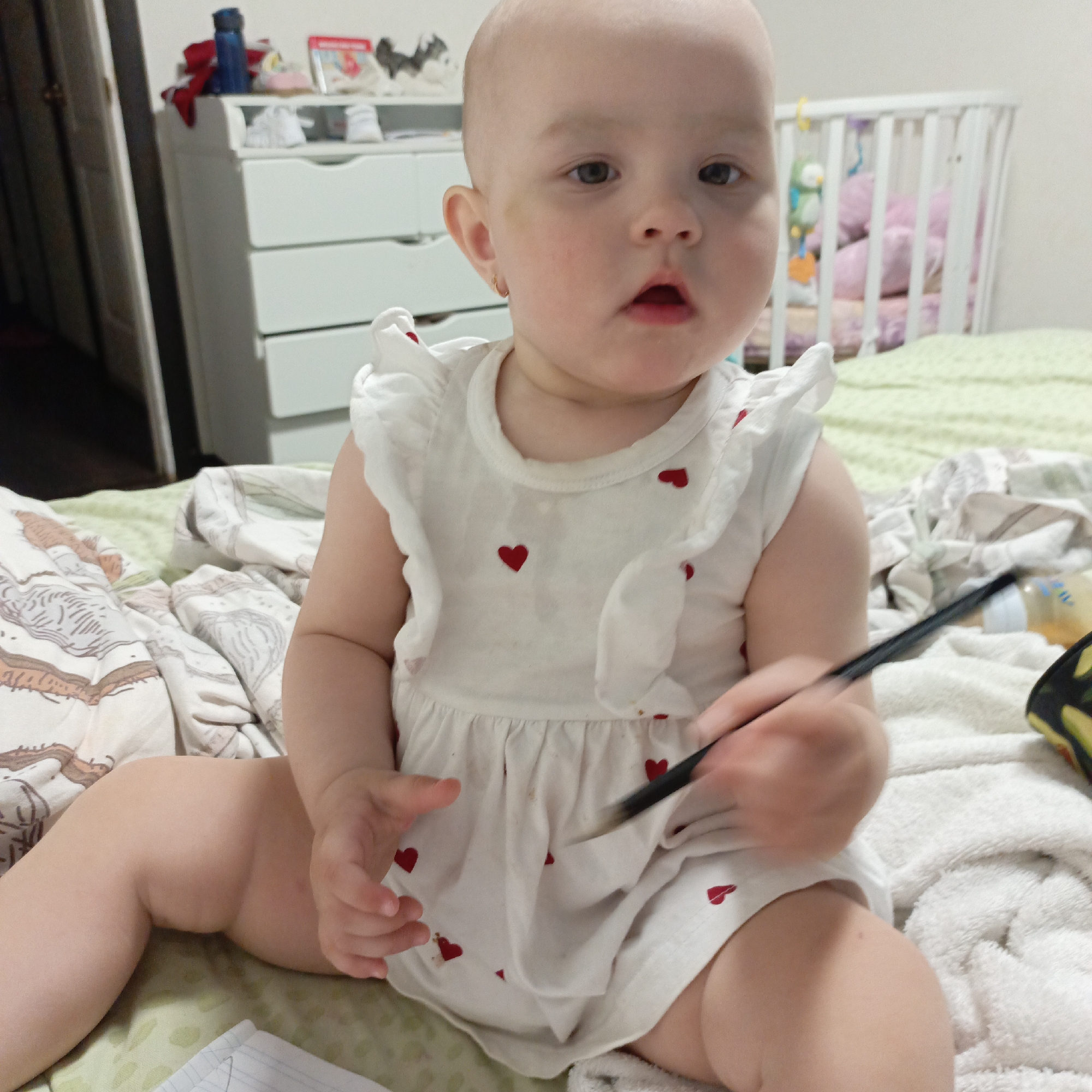 A fair-skinned baby is sitting on a bed in what appears to be a home setting in Unknown, Unknown. The baby is wearing a white dress adorned with red hearts and is holding a black pen in their right hand. The baby's expression is inquisitive, with eyes looking directly at the viewer.
The bed is covered with a mix of patterned and plain blankets. In the background, there is a white dresser with items on top and a white crib. A baby bottle and a case with a colorful pattern are visible to the right of the baby.