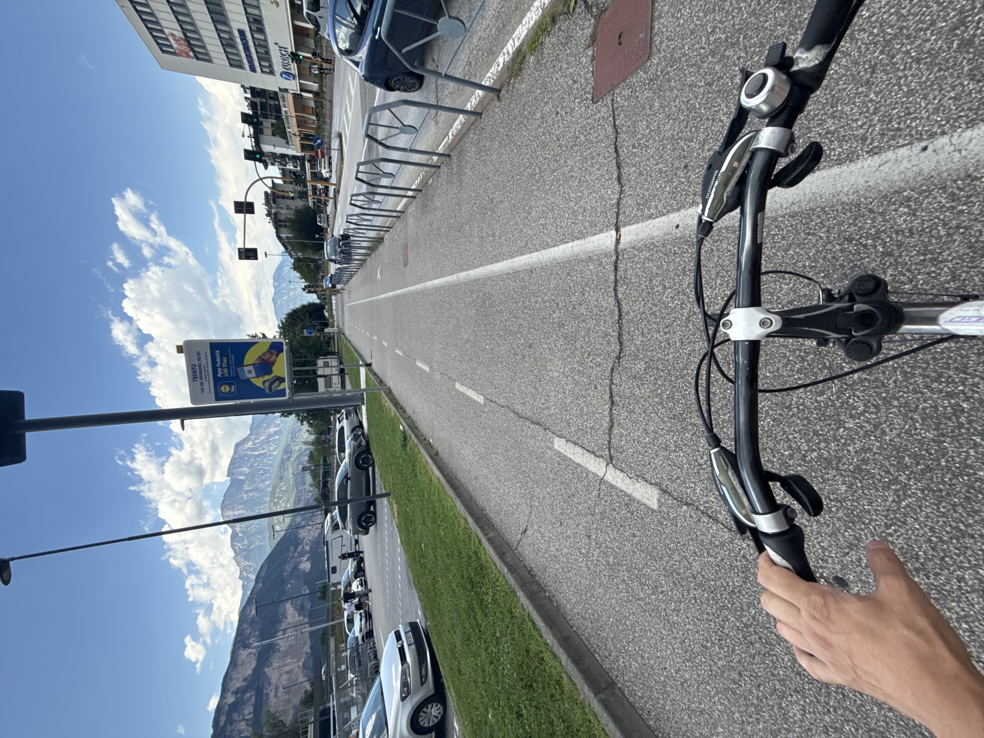 The image shows a first-person perspective of someone riding a bicycle on a dedicated bike lane in the city of Trento, Italy. The bike lane is paved and marked with white lines. To the left of the bike lane is a grassy area with a curb, and beyond that, a parking lot with several cars and a white RV. In the background, there are buildings, traffic lights, and a road with moving vehicles. Further in the distance, majestic mountains form a scenic backdrop under a blue sky with scattered clouds. A street sign is visible on the right side of the bike lane, which indicates "TRENTO VIA DEL BRENNERO, 99/101" and features a Lidl Plus advertisement. The handlebars of the bicycle are prominent in the foreground, with a hand gripping the left handlebar. The overall scene depicts a typical urban environment with natural elements and infrastructure for transportation.