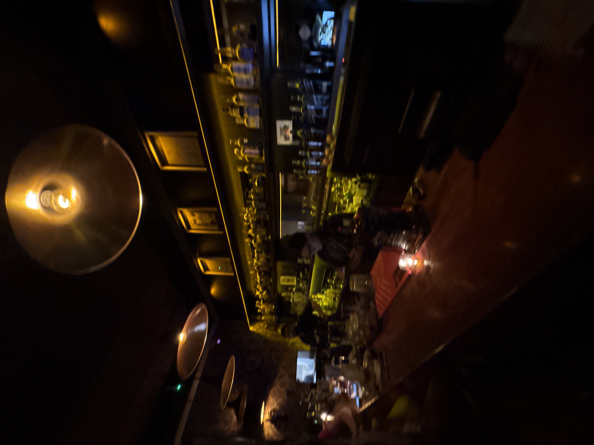The image depicts the interior of a bar in Mexico City, Mexico. The scene is dimly lit with prominent overhead lighting fixtures, including a large circular lamp on the left side of the frame. A bar runs along the right side, with bottles of alcohol displayed on shelves against the back wall. A bartender is working behind the bar. The overall atmosphere appears lively but dark.