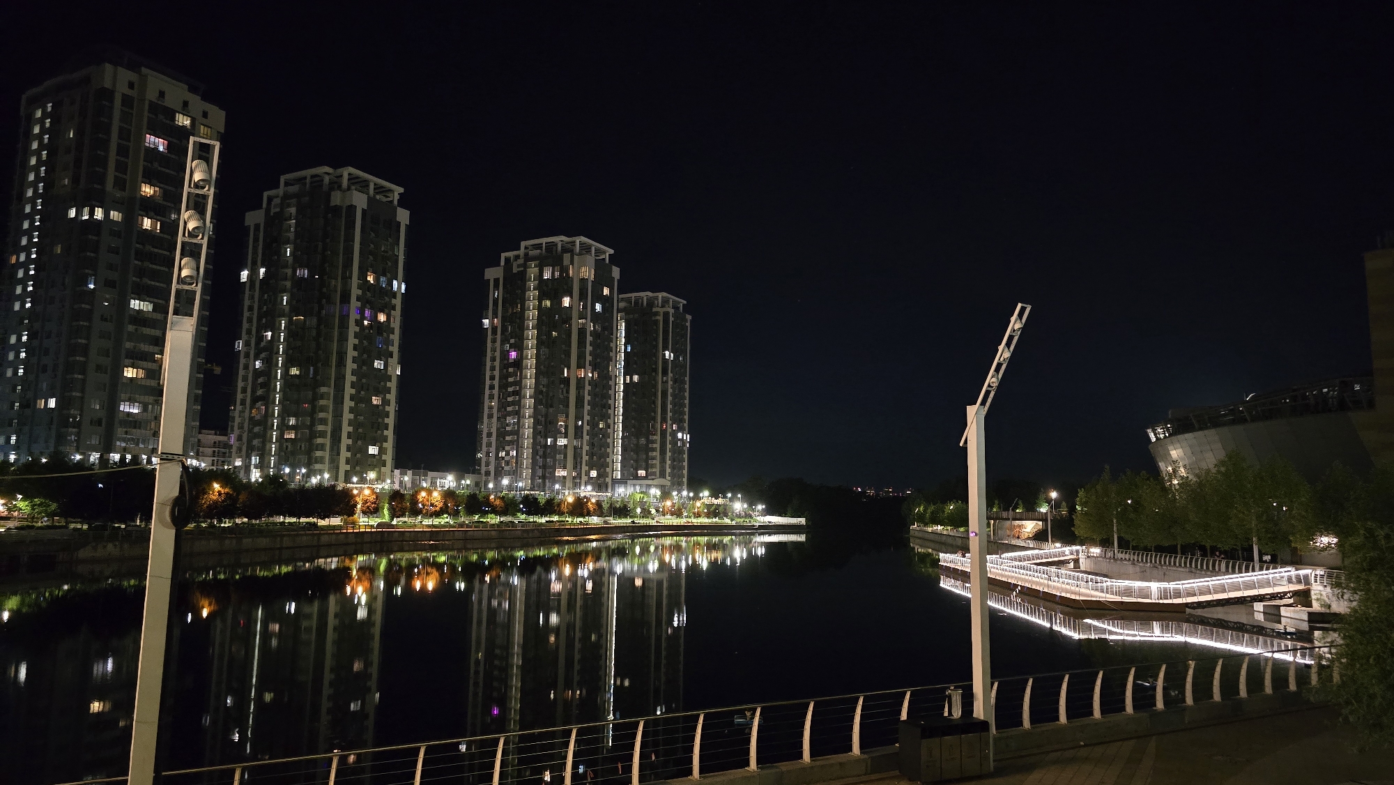 The image shows a night view of the Obolonska Naberezhna in Kyiv, Ukraine, featuring a row of modern high-rise residential buildings on the left. The buildings are illuminated with interior lights visible through the windows, casting reflections on the calm water of the Dnipro River in the foreground. A modern, white streetlamp stands prominently in the left foreground, its unusual design catching the eye. On the right side of the river, a contemporary, geometrically shaped building with a curved facade is visible, surrounded by trees. A boardwalk with decorative lighting runs along the waterfront, also reflecting in the water. The sky is dark, indicating nighttime, with a few faint stars visible. The overall scene is one of urban nighttime beauty, with a strong emphasis on architecture and its reflections in the water.
