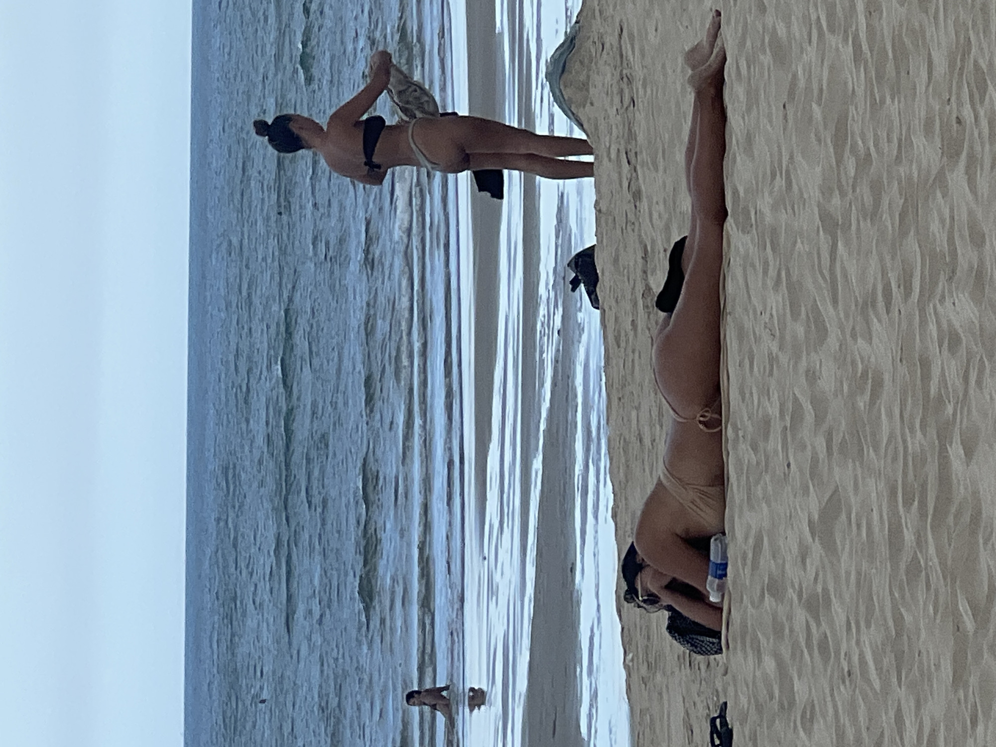 In Đà Nẵng, Vietnam, at a sandy beach with the ocean stretching to the horizon under a cloudy sky, two individuals are visible. In the foreground, a person lies prone on a tan towel, facing away from the camera, their head resting on a black bag, wearing a light brown bikini bottom. A white water bottle is placed beside them. Further back, near the water's edge, another person stands, facing away and towards the ocean, wearing a black bikini top and a light-colored bikini bottom. They are holding a rolled-up towel or garment. In the distance, a third person is seated in the shallow water. The beach exhibits distinct lines of wet and dry sand, with some debris and darker objects scattered near the waterline.