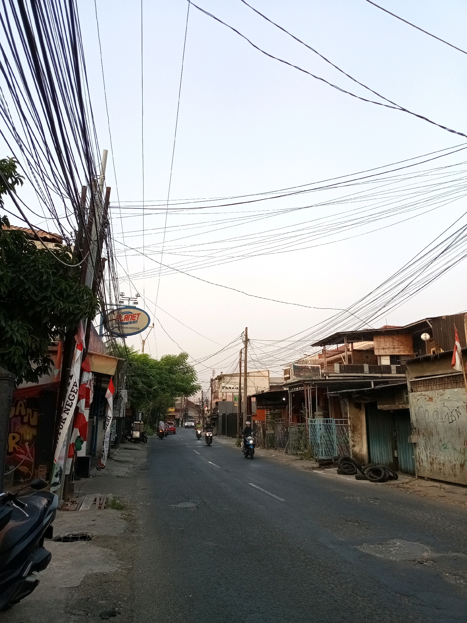 The image depicts a street scene in Bekasi, Indonesia, characterized by a dense network of overhead electrical wires crisscrossing the sky. The street is paved and appears to be a local thoroughfare with buildings lining both sides. On the left, there are shops with signage, including one that reads "PLANET BARI MOTOR." Several motorcycles are visible, with some in motion on the road and one parked on the sidewalk in the foreground. Indonesian flags are displayed on the left side of the street, suggesting a national or local event. The architecture of the buildings appears to be modest and functional, with some showing signs of wear and tear. The sky is clear and bright, indicating daytime, possibly late afternoon given the lighting. The overall impression is of a bustling, everyday urban environment in Bekasi.