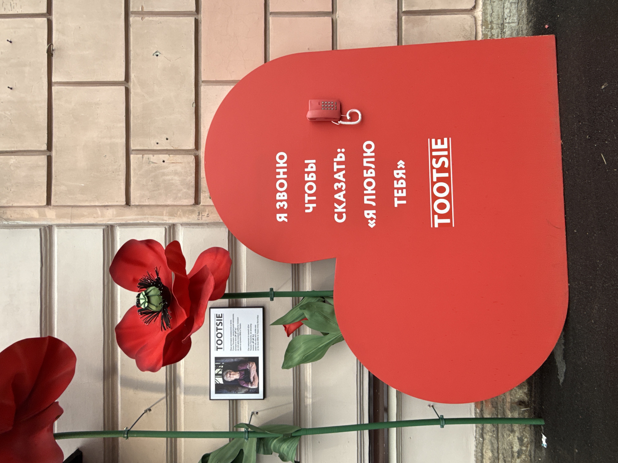 The image shows a red heart-shaped sign with the name "TOOTSIE" in white text. The sign also features a small red toy telephone and text in Russian, which translates to "I'm calling to say: I love you." A red poppy flower and a small poster with the TOOTSIE logo are placed next to the sign. The background includes a tiled wall and part of a street or sidewalk. The location is likely in Saint Petersburg, Russia, based on the presence of the Cyrillic alphabet.