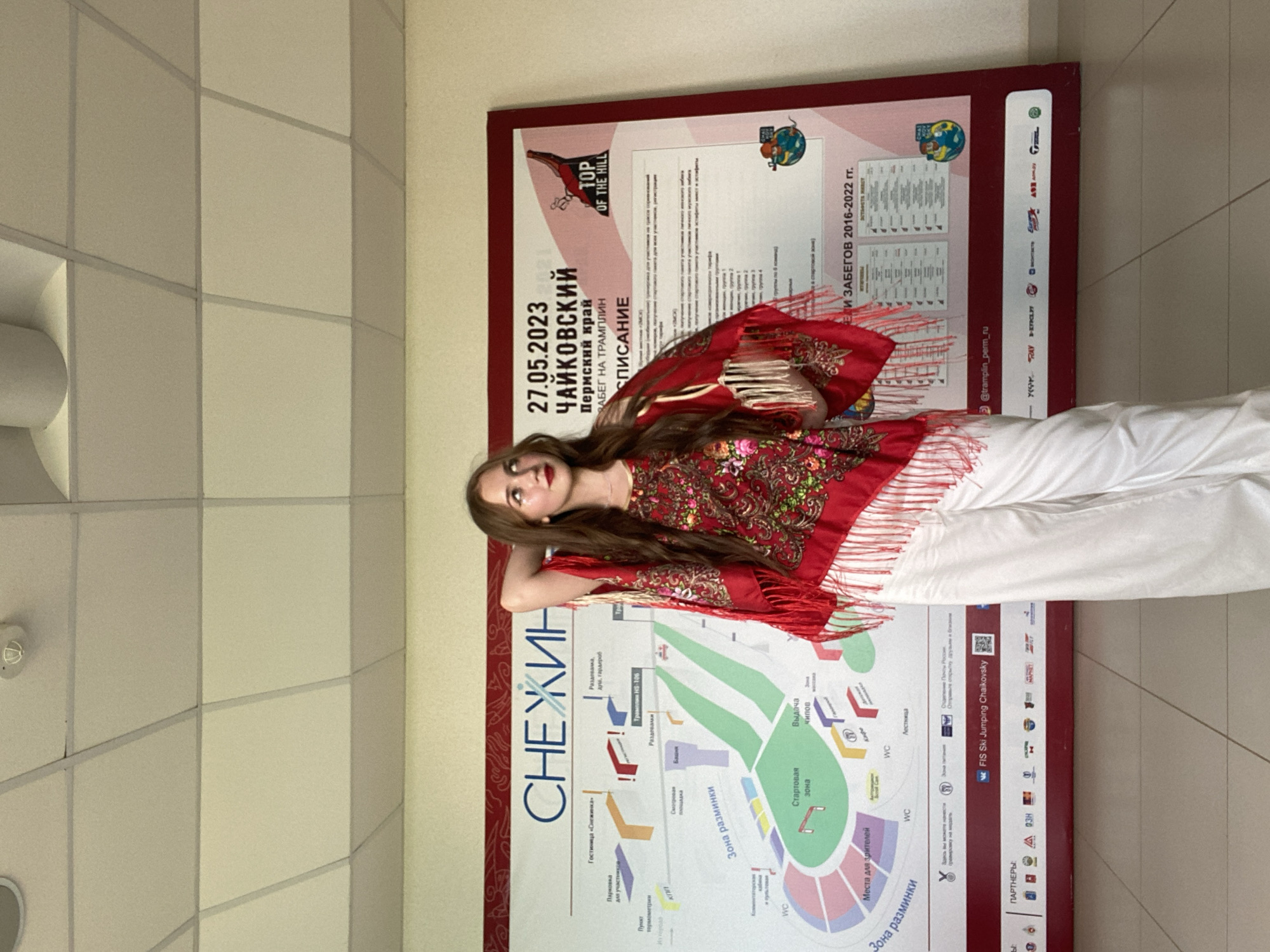 A young Caucasian woman with long, auburn hair and fair skin stands in front of a large informational poster. She is wearing a red shawl with colorful floral embroidery and white fringe, and loose white pants. Her right arm is bent behind her head, and she is looking up and to her left. The poster displays a map of a ski jumping complex, with titles in Russian. The top of the poster reads "27.05.2023 ЧАЙКОВСКИЙ Пермский край ЗАБЕГ НА TRAMPLIN" which translates to "27.05.2023 Chaikovsky Perm Krai Trampoline Competition." The location appears to be an indoor venue, indicated by the tiled ceiling and fluorescent lighting. The overall scene suggests an event related to winter sports in the Perm Krai region of Russia.