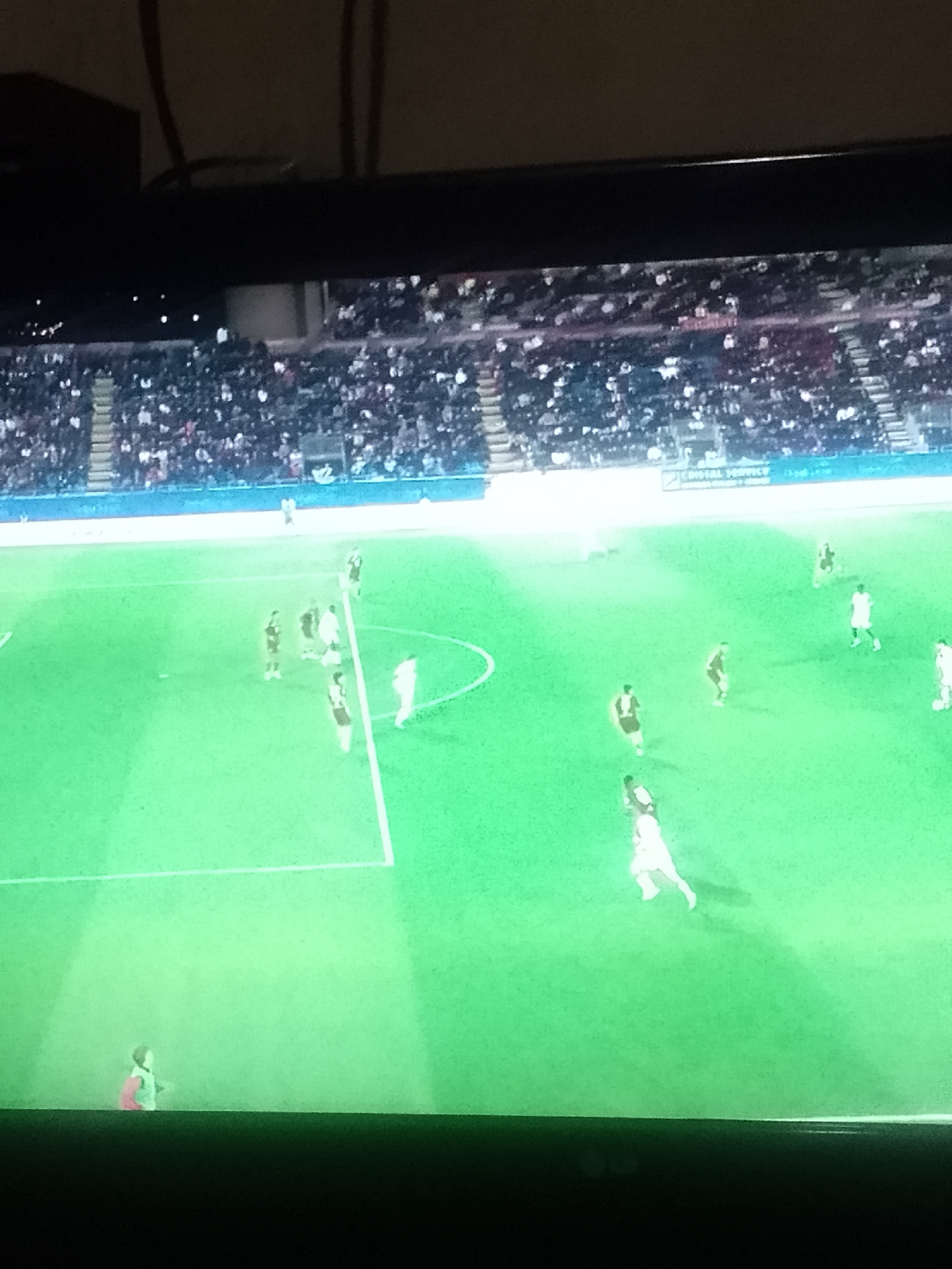 A television screen displays a soccer game in progress. The playing field is green with white lines marking boundaries. Multiple players are visible, some in white uniforms and some in dark-colored uniforms, engaged in active play. A stadium with a large crowd of spectators surrounds the field. The location is Unknown, Unknown.