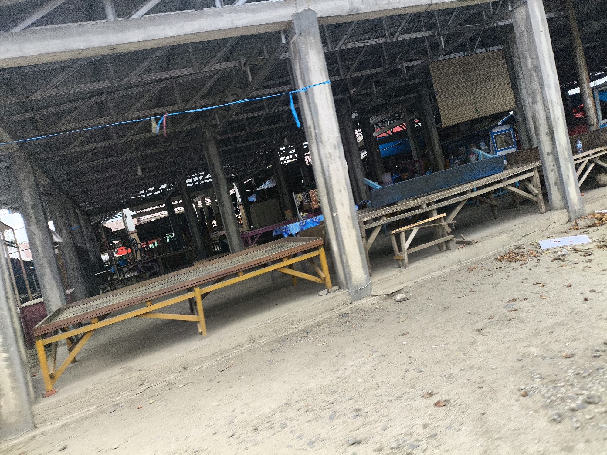 The image depicts an outdoor market structure with a corrugated metal roof supported by concrete pillars. Several wooden benches or tables are present. One prominent yellow-legged bench is in the foreground. In the background, there are stalls with blue tarpaulins and a woven bamboo blind. A blue cord with colorful tags is strung across the ceiling. The ground is sandy and appears to be a mix of sand and gravel, with some scattered debris, including a white sheet of paper and some small round objects. The scene is static, suggesting the market may be closed or in a period of low activity. No people are clearly visible in the frame. The environment appears to be an open-air market in Harapan, Indonesia.