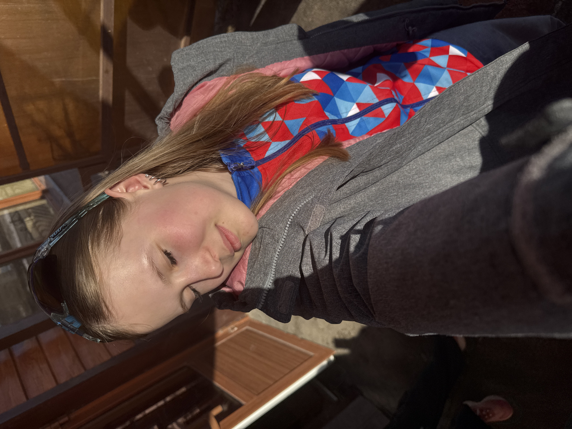 A woman is shown in an outdoor setting with natural light. She is wearing sunglasses on her head and has multiple earrings in her left ear. She is wearing a grey jacket over a red and blue jacket. She is outside, possibly in a sunny area near a wooden structure. There's no clear indication that this image was taken in Gory, Russia.