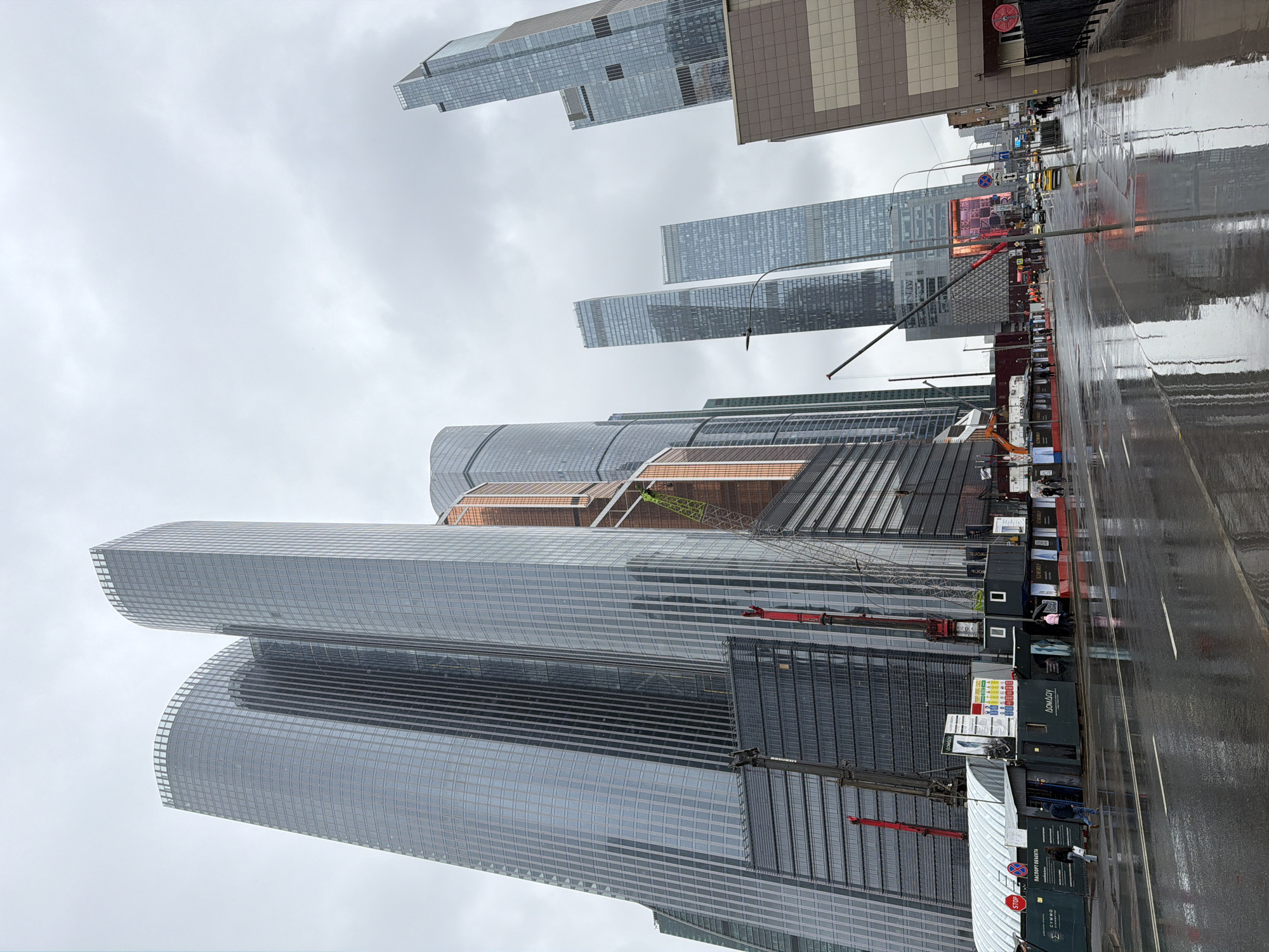 The image depicts a modern cityscape under an overcast sky, likely in Moscow, Russia, given the context. The scene is dominated by numerous tall, contemporary skyscrapers, primarily constructed from glass and steel, reflecting the dull, cloudy sky.
In the foreground and center, there are two prominent, massive glass skyscrapers with distinctive curved facades, suggesting a high-tech architectural style. To their right and further back, several other tall, rectangular glass towers rise into the sky, creating a dense urban environment. One of the buildings in the mid-ground features a visible structure with exposed brown/copper-colored elements and scaffolding, indicating ongoing construction or an unfinished section.
The ground level features a wide, multi-lane street that appears heavily wet, reflecting the surrounding buildings and suggesting recent or ongoing rain. White road markings are visible on the asphalt. Along the right side of the street, there's an active construction site. It is lined with numerous dark-colored modular containers or temporary buildings. Several cranes are visible: a large green lattice boom crane positioned among the taller buildings, and multiple smaller red and black mobile cranes near the construction containers. An orange excavator can also be seen at the construction site.
Vehicles, including several cars and at least one yellow taxi, are present on the wet road, some with headlights on, indicating movement. A few pedestrians are visible walking on the sidewalk next to the construction site.
Visible text includes a red octagonal "СТОП" sign (Russian for "STOP"), a blue sign with "ВХОД" (Entrance), and several signs on the construction containers or advertising boards with Russian text, such as "ДОМОДЕ" and "ВНУКОВО" (likely referring to Moscow airports), and "ДОМАОУ". "DOPAY" is also visible on one of the containers. The overall impression is a bustling urban area with significant modern development and ongoing construction, under typical grey weather conditions.