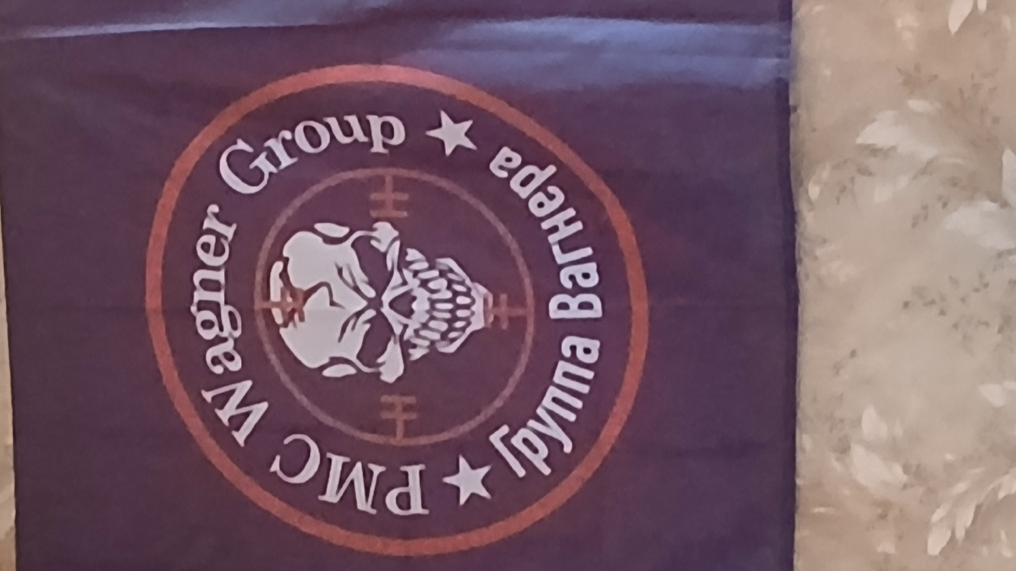 The image shows a dark purple flag with a circular emblem. The emblem features a stylized skull in the center, superimposed on what appears to be a reticle or target. Text in white lettering encircles the skull. On the upper half, it reads "PMC Wagner Group", and on the lower half, it reads "Группа Вагнера". The text is separated by white stars on either side. The flag is hanging and partially overlaps a textured surface that appears to be wallpaper with a floral pattern. The lighting suggests an indoor setting. There is no visible indication of the city of Mozhaisk, Russia.