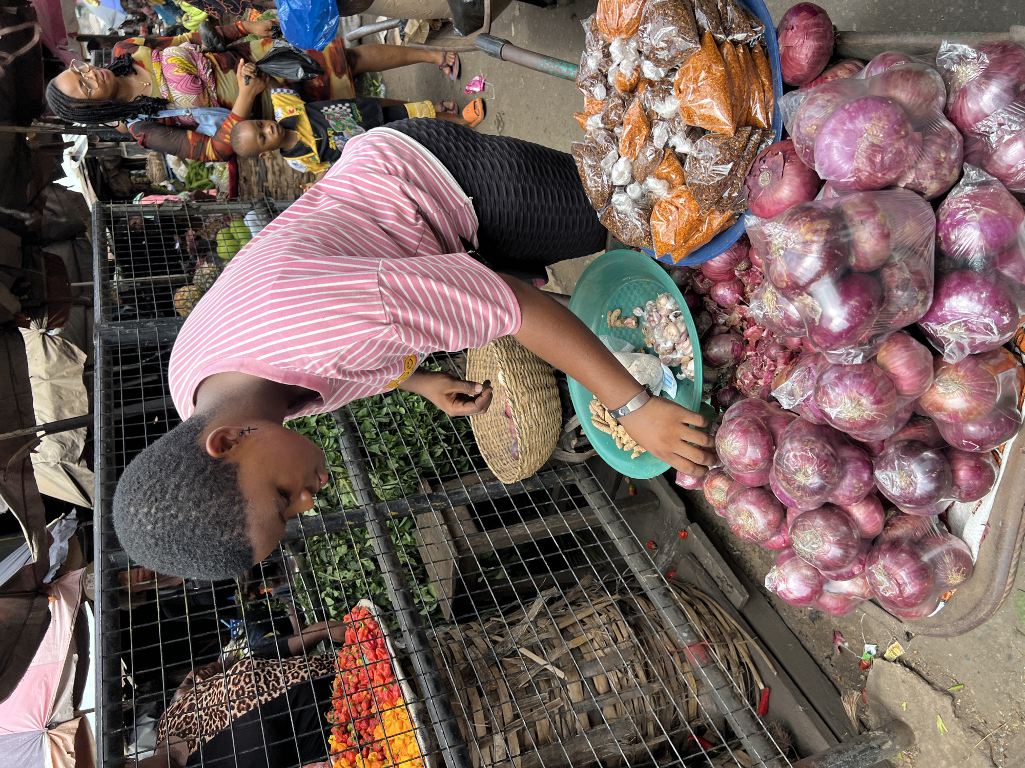 The image, taken in Nkpor, Nigeria, depicts a market scene. A person wearing a pink and white striped shirt and a black skirt is bent over a metal grate, sorting produce. A bowl of garlic and spices in plastic bags are visible nearby. To the right, there are piles of red onions, some wrapped in plastic. Under the grate, red and yellow chilies are visible. In the background, an adult and a child are lying down on some merchandise or textiles. The ground appears to be concrete.
