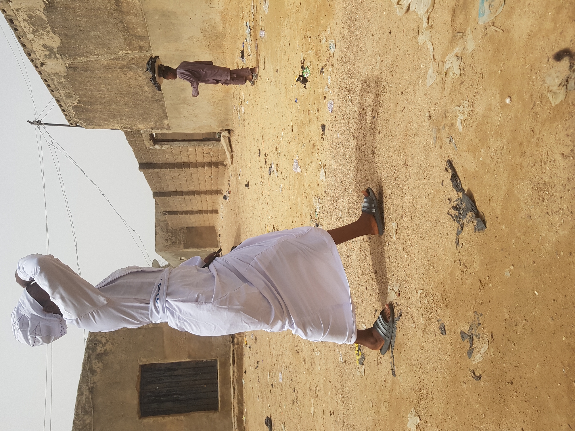 A medium shot captures two individuals in Dutsinma B, Nigeria. The foreground features a man facing away, clad in a white robe and head covering, standing on a dirt ground with scattered litter. He appears to be adjusting his head covering with both hands. In the background, another person in a long-sleeved maroon outfit and a straw hat, standing on the same dirt ground. The environment includes dilapidated structures on both sides, and the sky is overcast.
