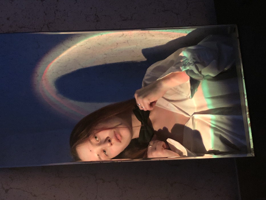 This image depicts a reflection of a young female in a rectangular mirror, observed in an indoor setting in the city of Фоки, Russia. The female has long, light brown hair and a fair complexion, with a small dark mole or spot visible on her forehead. She is wearing a light-colored, possibly white or cream, long-sleeved collared shirt, unbuttoned at the neckline. A dark-colored bow tie is fastened around her neck, which she is adjusting with both hands. Rings are visible on the ring fingers of both hands. A prominent, arcing light projection displaying rainbow colors (red, orange, yellow, green, blue, purple) illuminates the scene, appearing on the wall to the right of the mirror and reflected within the mirror itself. The background within the mirror behind the subject appears dark blue or purple, likely influenced by the light projection. The overall lighting is low, with the colored light source providing key illumination and casting distinct color washes across the subject and her attire.