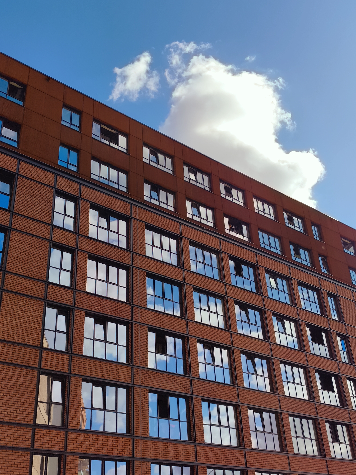 This is a low-angle, full-frame, vertical shot of a modern, multi-story building with a brick facade and a burnt orange-colored upper section. The building features numerous windows with dark frames, many of which reflect a bright blue sky with scattered white clouds. The architecture suggests an urban setting, possibly an apartment complex or office building. The scene is devoid of any people or vehicles, focusing solely on the structure and its reflection of the environment. The image is sharp and well-lit, with vibrant colors. While the image itself does not contain explicit indicators of its location, the request specifies it is in Gdańsk, Poland.