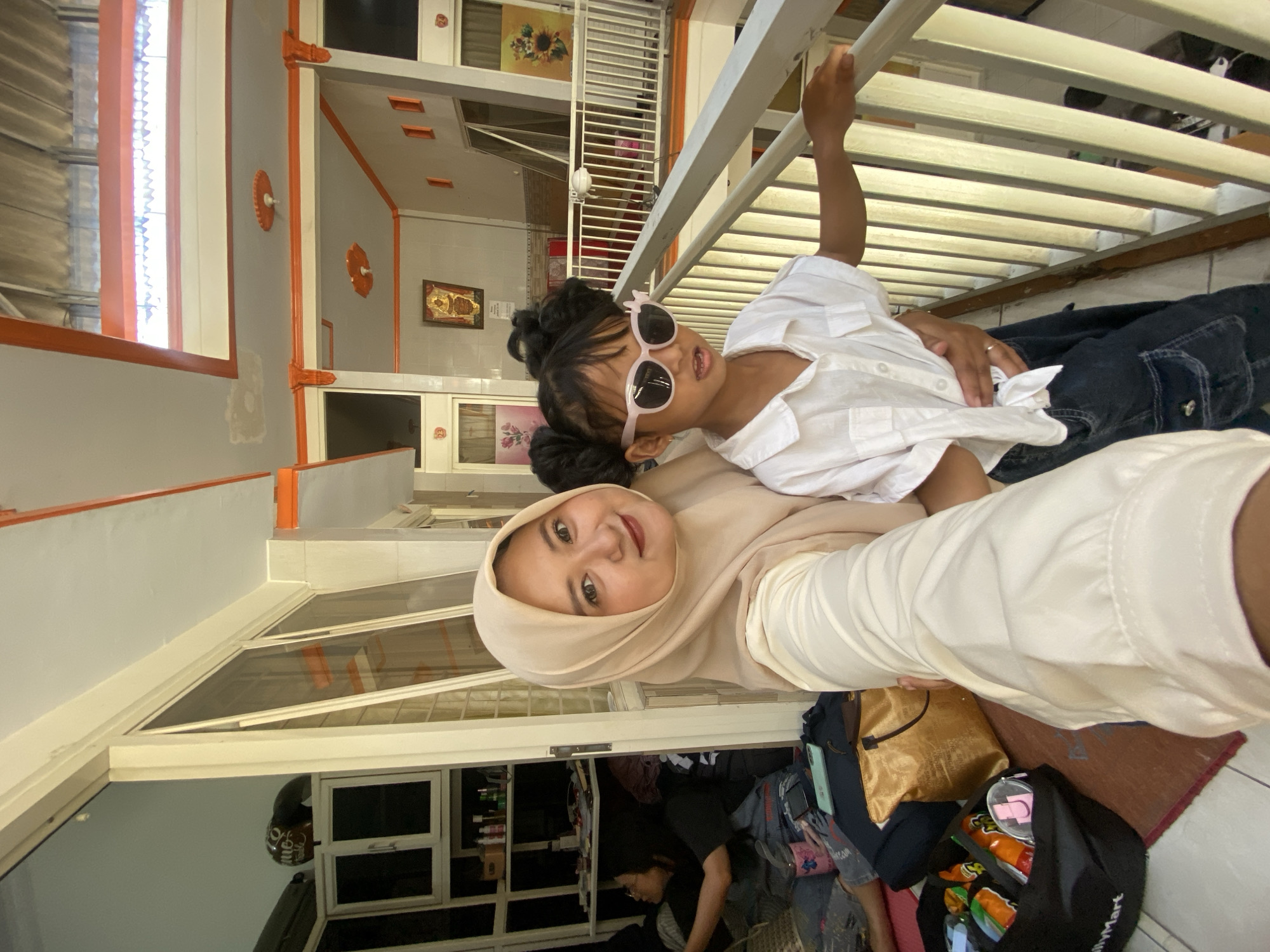 A woman wearing a hijab holds a child wearing sunglasses as she takes a selfie. The subjects are on the balcony or stoop of a home in Jember, Indonesia. The woman is wearing a light-colored top and tan hijab. The child has pigtails and a white shirt. Behind them is a white railing and a multi-story building with decorative elements painted in orange. Below them are the contents of a bag, including various snacks in their original packaging. Another person, seemingly working in the home, is visible in the background.