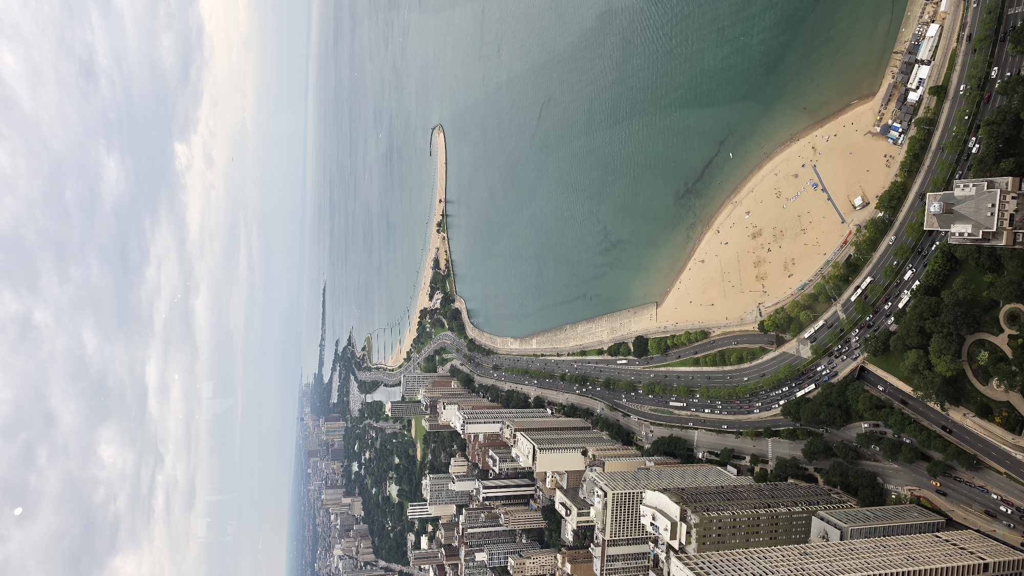This is an aerial photograph taken in Chicago, Illinois, United States of America, looking down at a panoramic view of the city's lakefront. The image captures a vast expanse of Lake Michigan on the right side, its waters a deep teal color under an overcast sky. Along the shoreline, a curve of sandy beach is visible, populated by a scattering of sunbathers and beachgoers. A concrete promenade runs along the edge of the beach.
To the left of the lakefront, a multi-lane highway, likely Lake Shore Drive, is densely packed with traffic, including numerous cars and buses. This highway is bordered by a strip of lush green parkland, featuring numerous trees. Beyond the park and highway, the cityscape rises with a dense concentration of tall, modern buildings, including skyscrapers and apartment complexes, indicative of a major urban center. The buildings vary in height and architectural style, with many featuring glass facades and repeating patterns of windows. The overall scene depicts a bustling urban environment integrated with a natural shoreline and recreational area.