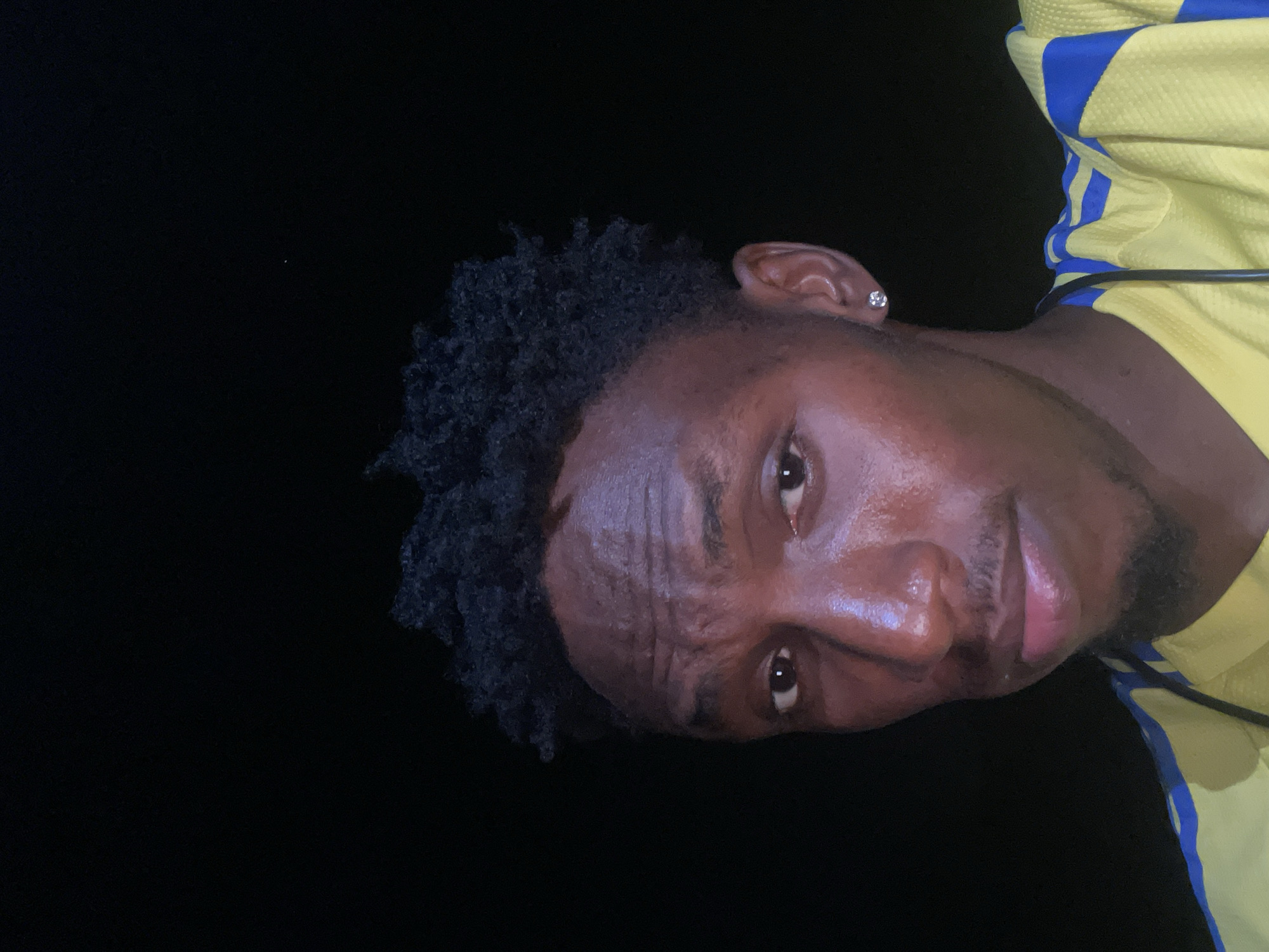 A close-up portrait of a Black man with dark, curly hair and a small earring in his left ear. He is wearing a yellow shirt with blue stripes. The background is black, suggesting a night setting.