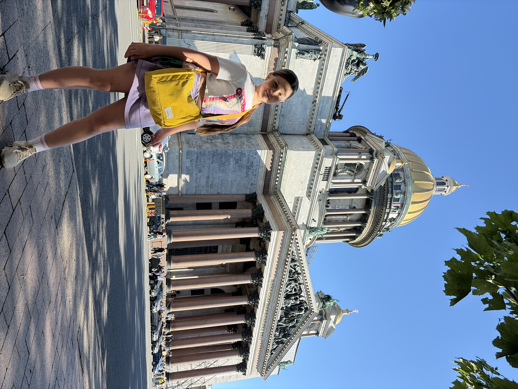 A young woman stands in front of St. Isaac's Cathedral in Saint Petersburg, Russia. She is wearing a white crop top with a graphic print, a purple skirt, white socks with pink stripes, and beige sneakers. She has a yellow crossbody bag slung over her left shoulder. The cathedral is a large, imposing structure with a golden dome, classical columns, and intricate sculptures. The sky is clear and blue, and sunlight casts shadows on the pavement. Several cars are parked on the street in front of the cathedral. The woman is looking towards the camera with a slight smile. The scene is captured from a low angle, emphasizing the grandeur of the cathedral.