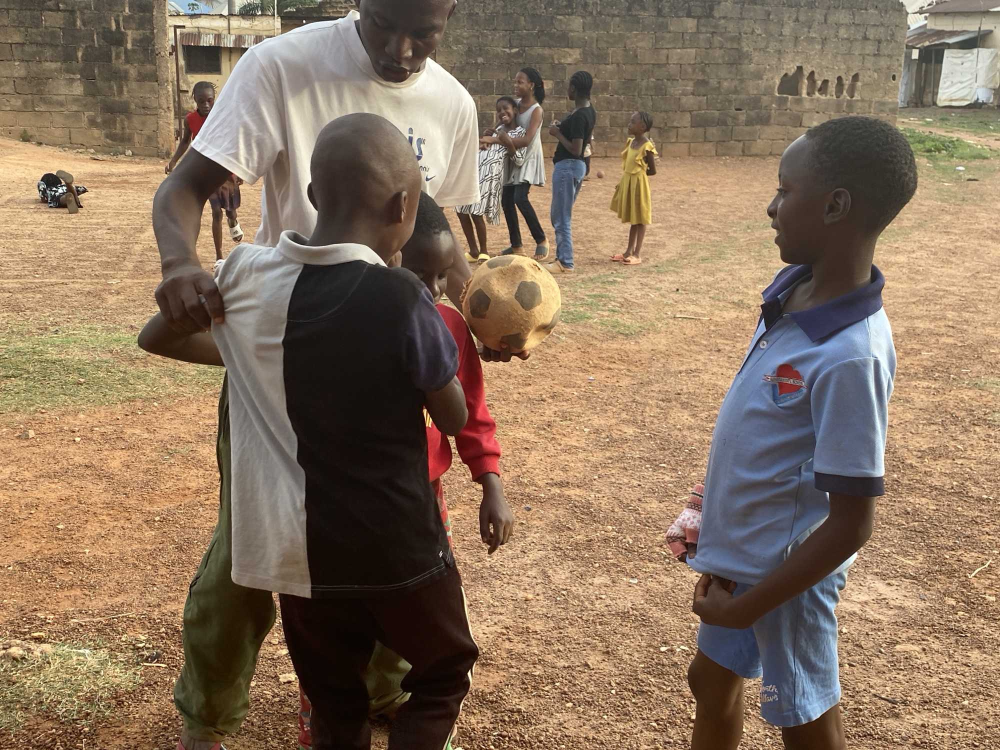 An outdoor scene in Bukuru, Nigeria, captures an active moment among children and an adult on a dusty, light-brown ground. In the foreground, an adult male wearing a white t-shirt with a visible "NIKE" logo looks down, interacting with three children. One child, seen from behind in a black and white polo shirt, has their hand on the adult's arm. Another child, partially visible in a red long-sleeved top, holds a well-worn, light-colored soccer ball. Facing them from the right is a child in a light blue polo shirt and matching shorts, which have "Growth Love" printed on them; their shirt also features a small, heart-shaped logo.
In the midground, a group of three individuals – two women and a child – stand to the left of the adult, engaged in conversation or observing, with one woman smiling. Further back, a young girl in a yellow dress stands to the right, looking towards the left. To the far left in the background, a child in red is seen lying down on the ground. The background features a brick wall, partially damaged, behind which simple structures with corrugated metal roofs are visible, suggesting a community or village setting. The overall impression is one of communal activity, possibly involving a game of soccer, under a clear, bright sky, likely in the late afternoon.