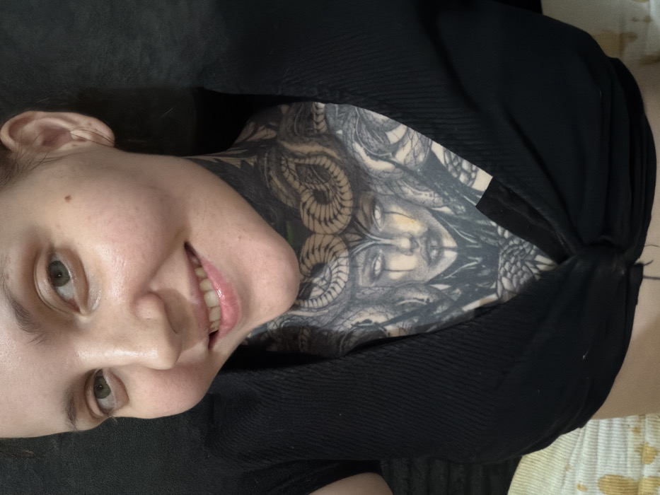 A woman lies on her back, smiling. Her face is angled towards the left side of the frame. She has fair skin, green eyes, and light brown hair. She wears a black top that partially covers her chest and neck. A detailed black and gray tattoo covers her chest and neck. The tattoo appears to depict a woman’s face surrounded by snakes. The background is slightly blurred and indistinct.