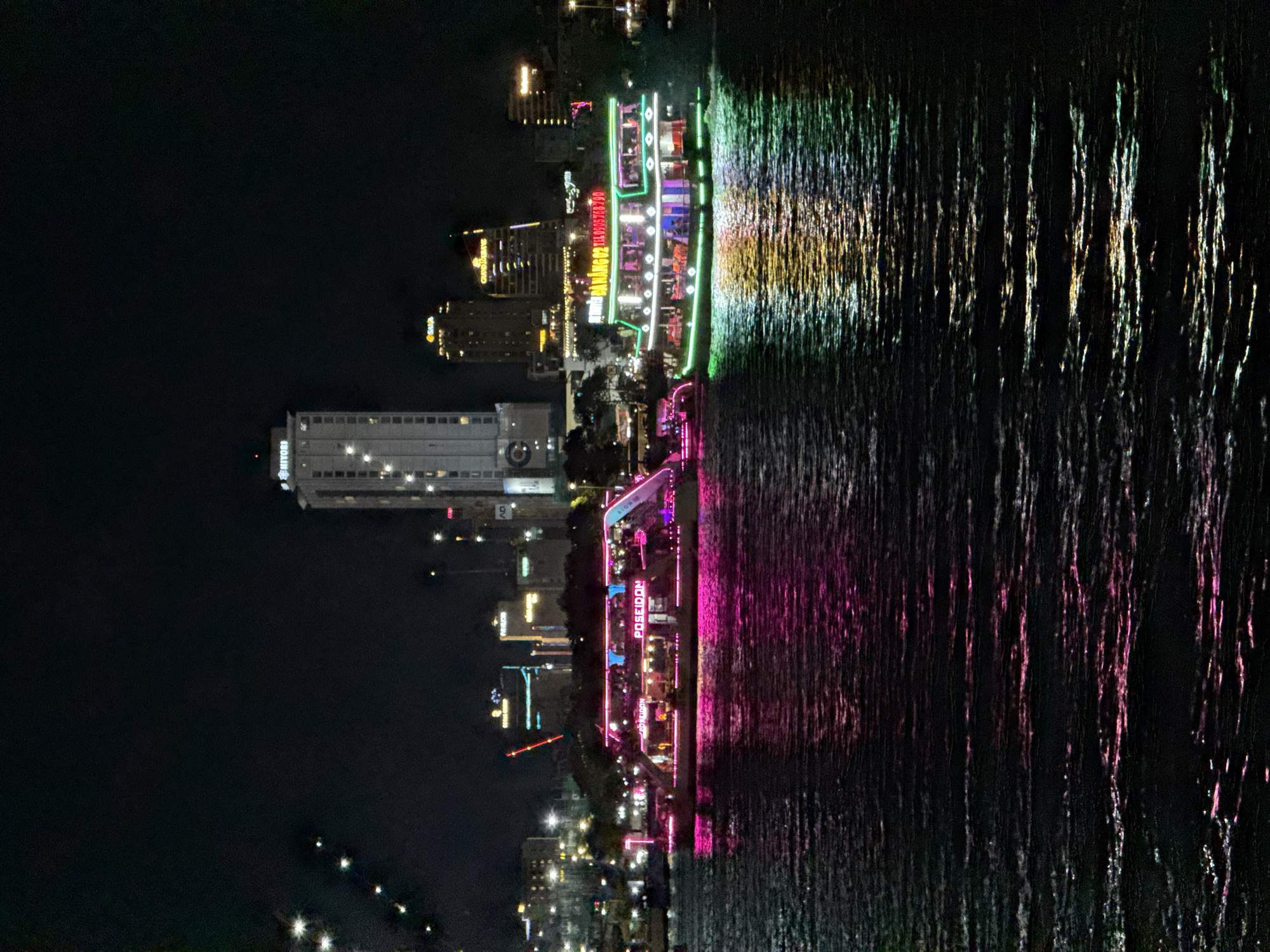 This is a nighttime photograph of the Han River in Da Nang, Vietnam. Two illuminated tour boats are the main subjects, docked or moving slowly on the water. The boat on the left is adorned with vibrant pink lights and has "POSEIDON" prominently displayed on its side. The boat on the right is lit with green and white lights and has text that appears to be in Vietnamese, including "ĐÀ NẴNG 02" and a phone number.
In the background, the Da Nang skyline is visible, featuring several tall buildings illuminated at night. One prominent building on the left is a modern, white high-rise with the "Hiyori" logo at its peak. Other buildings exhibit various illuminated signs and windows. The river's surface reflects the colorful lights from the boats and the city, creating shimmering patterns on the dark water. The overall atmosphere is lively and urban, characteristic of a popular tourist destination at night.