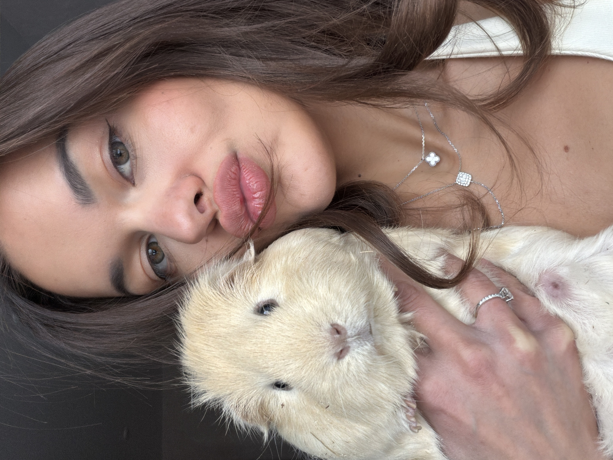 A woman with long brown hair, wearing a silver necklace with clover and square pendants and a ring on her left ring finger, is laying down and holding a light beige guinea pig. The location in the city of Krasnoyarsk, Russia is not discernible from the image.