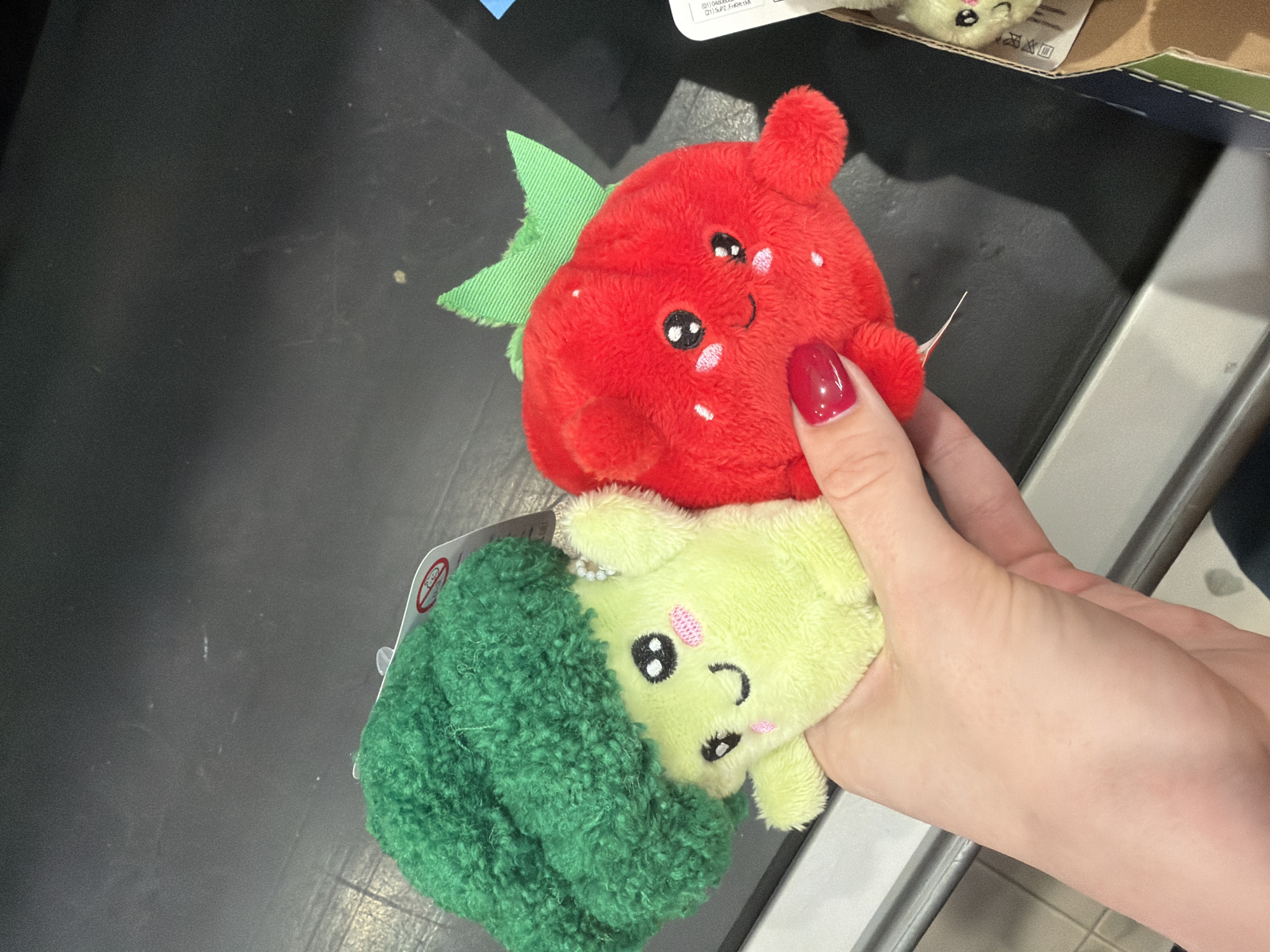 A person's hand holds two plush toys against a dark, possibly black, surface, likely a store counter or conveyor belt, in Berezniki, Russia. The hand, with red nail polish on the thumb and index finger, is fair-skinned. One plush toy is a bright red strawberry with a green leaf at its top. It has black eyes, a small smiling mouth, and pink blush marks on its cheeks, along with white embroidered 'seeds' on its body. The second plush toy is a light green, soft-textured broccoli head, with a darker green, more textured 'stem' or florets wrapping around it. Like the strawberry, it features black eyes, a small smiling mouth, and pink blush. Both toys appear to be part of a set of cute, anthropomorphic fruit and vegetable plushies.
In the background, to the upper right of the red strawberry, a white product label with black text and symbols is partially visible. Legible portions include "(01) 062020", "(21) 3JPS", and "Fraberik". A white tag with a red circle-slash symbol (often indicating a safety warning) is also partially visible near the green broccoli. The setting appears to be indoors, possibly a retail environment, given the presence of product labels and the type of counter. The overall impression is one of playful, soft toys being presented or purchased.