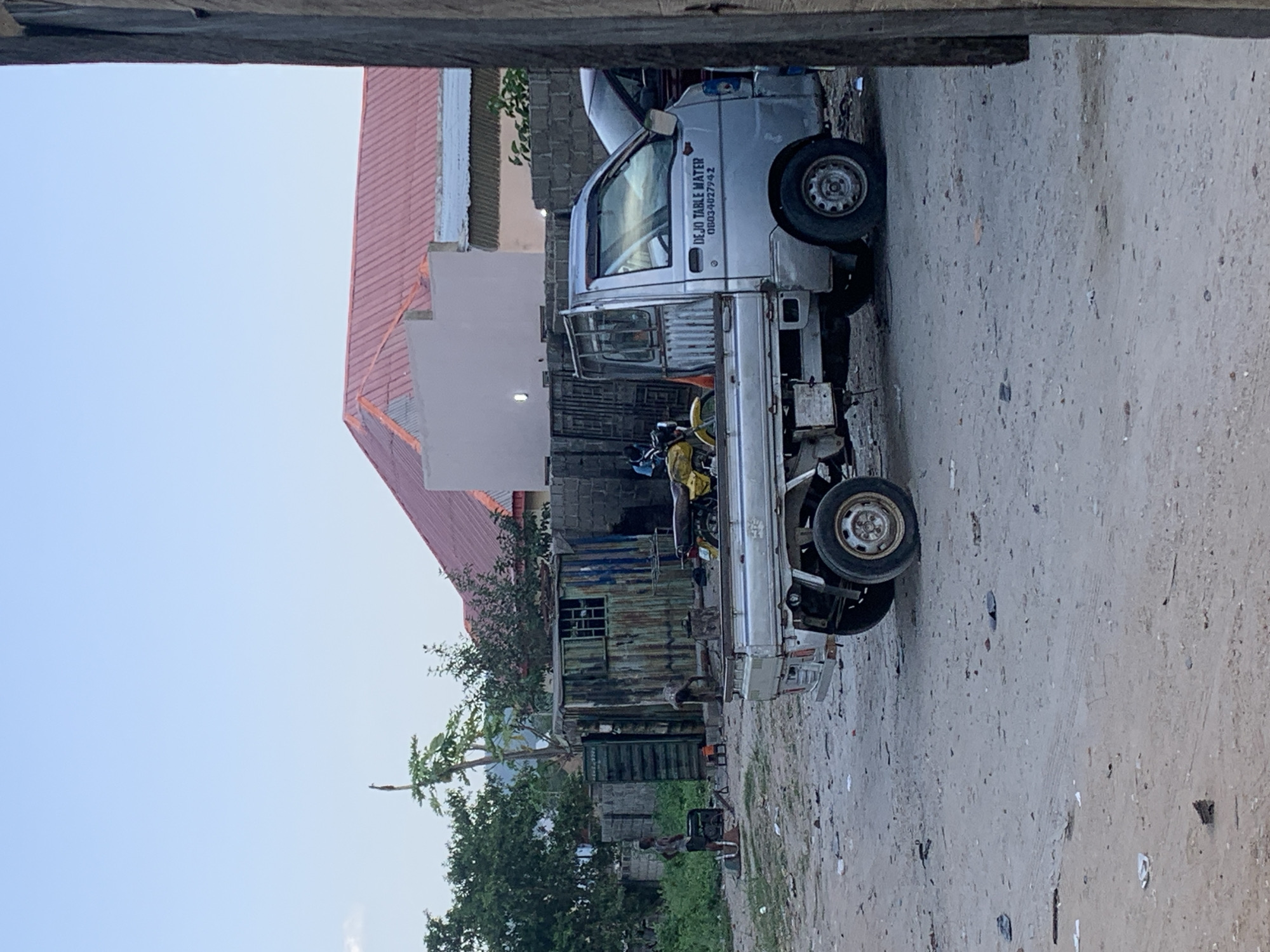A silver mini-truck is parked on a dirt road in Adankolo, Nigeria. The truck displays the text "DEJO TABLE WATER" along with a phone number. The surroundings include a partially visible building with a red roof and a small makeshift structure made of corrugated metal and wood. Green vegetation is also present. There are at least two people in the scene, and what appears to be a motorcycle is in the bed of the mini truck. The sky is visible above the structures and trees.