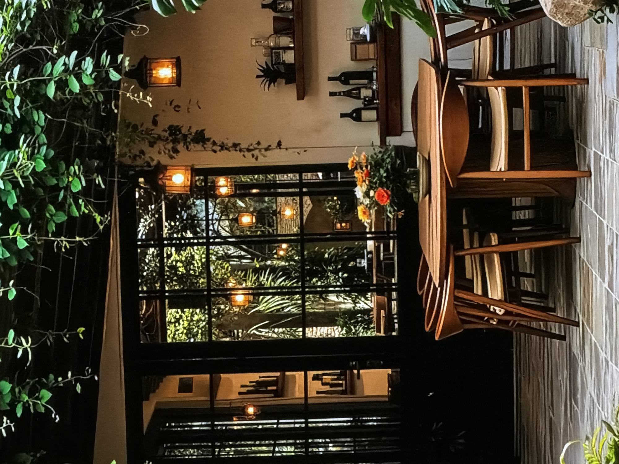 The image displays an inviting interior space, likely a restaurant or bar, characterized by warm, atmospheric lighting and lush greenery. The scene is oriented vertically in the provided image; when corrected, it depicts a cozy, evening ambiance.
In the foreground, vibrant green foliage, possibly vines, cascades down the left side, partially obscuring a light-colored wall. A single wall-mounted lantern with a warm orange glow illuminates this area. Further into the room, a large, dark-framed window or glass partition divides the view. Through this window, more greenery is visible, interspersed with several glowing lanterns, suggesting an outdoor garden or courtyard area.
To the right of the window, against a light-colored wall, are two wooden shelves. These shelves hold various bottles, likely wine or spirits, along with glassware and a black artificial pineapple as decoration. Below the shelves, near the window, a vase holds a colorful bouquet of orange and yellow flowers.
On the far right, a stack of four wooden chairs is visible, their curved designs and warm wood tones contributing to the room's aesthetic. These chairs appear to be neatly stacked, possibly indicating the establishment is closed or preparing to close. The wall behind the chairs features a textured, possibly tiled, surface.
The overall impression is one of tranquility and warmth, set during the evening or night, with no people present. There is no visible text. The setting suggests a sophisticated and comfortable establishment in Sofiivska Borshchahivka, Ukraine.