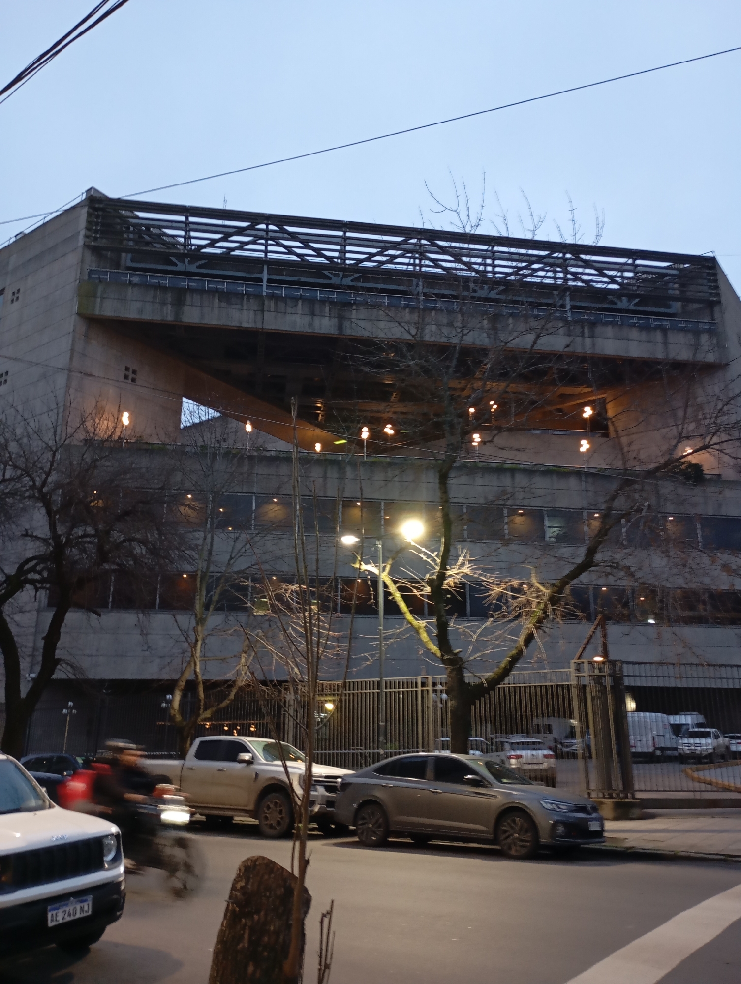 The image shows a grey, modern building with exposed concrete and a metal structure on its upper levels. The building is set against a twilight sky. Several bare trees are visible in front of the building, their branches reaching upwards. A street with a sidewalk runs in front of the building, and several vehicles are parked or driving on the street. One vehicle is a white pickup truck, another is a silver sedan, and a white SUV is partially visible on the left. A person on a motorcycle, blurred due to motion, is riding on the street. String lights with warm illumination are strung across the upper levels of the building. The environment suggests an urban setting, likely in La Plata, Argentina, given the license plate on the white SUV which reads "AE 240 NJ". The overall scene depicts a street view in front of a large building during the evening.