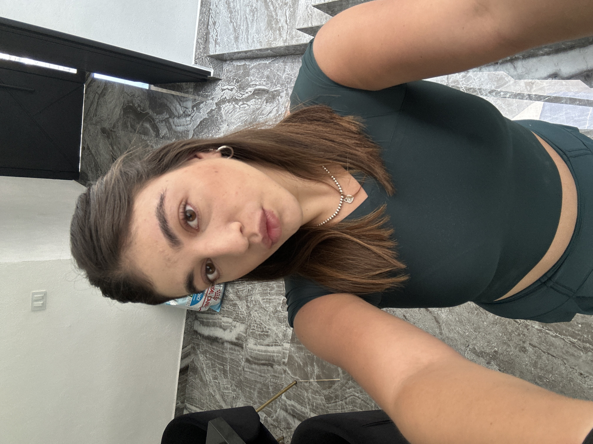 A young woman with brown hair and eyes is positioned in a selfie taken indoors. She is wearing a dark green athletic top and matching leggings, with a silver necklace and hoop earring. The background features a patterned marble floor and wall, a white wall with a light switch, and a black metal gate. There is a bag of Royal Canin dog food visible. A portion of black chairs is present at the bottom of the image. The location is in Puebla, Mexico, based on the Royal Canin bag, which is a brand popular in that region.