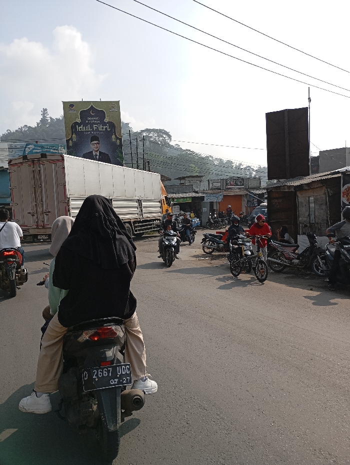 The image shows a street scene in Kutawaringin, Indonesia, with a billboard in the background. The billboard reads "Selamat Hari Raya Idul Fitri 1443 H" and features an image of a man in traditional clothing. In the foreground, there are several people riding motorbikes. There are also several motorbikes parked on the side of the road. The road is paved and there are power lines overhead. There are several buildings in the background, including a large building with a flat roof, a smaller building with a corrugated metal roof, and a building with a wooden facade. The image is taken during the day with clear skies. The scene is a typical street scene in Kutawaringin, Indonesia.